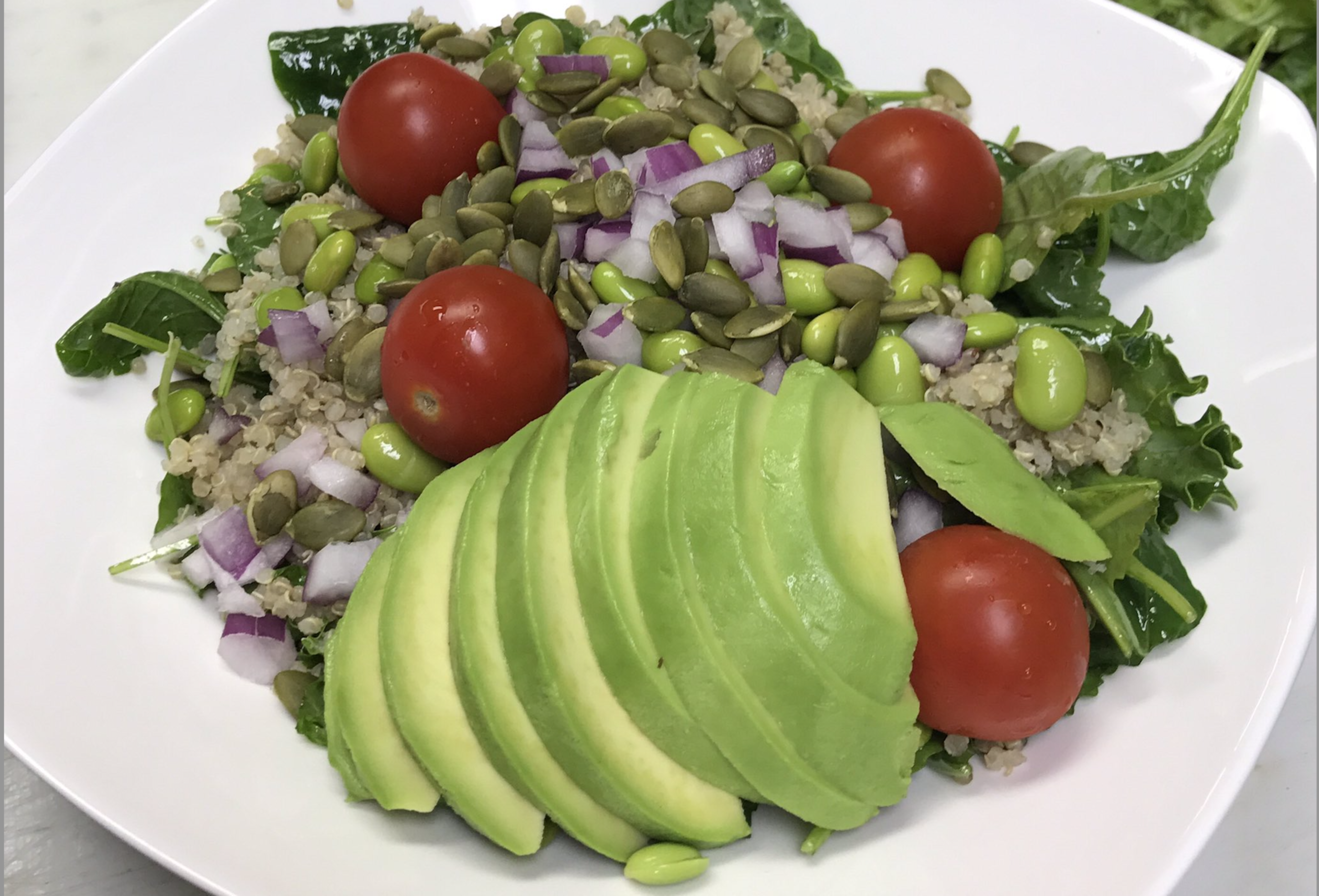 Sliced avocado, cherry tomatoes, chopped red onion, pumpkin seeds, edamame, quinoa, and leafy greens are arranged on a white plate.