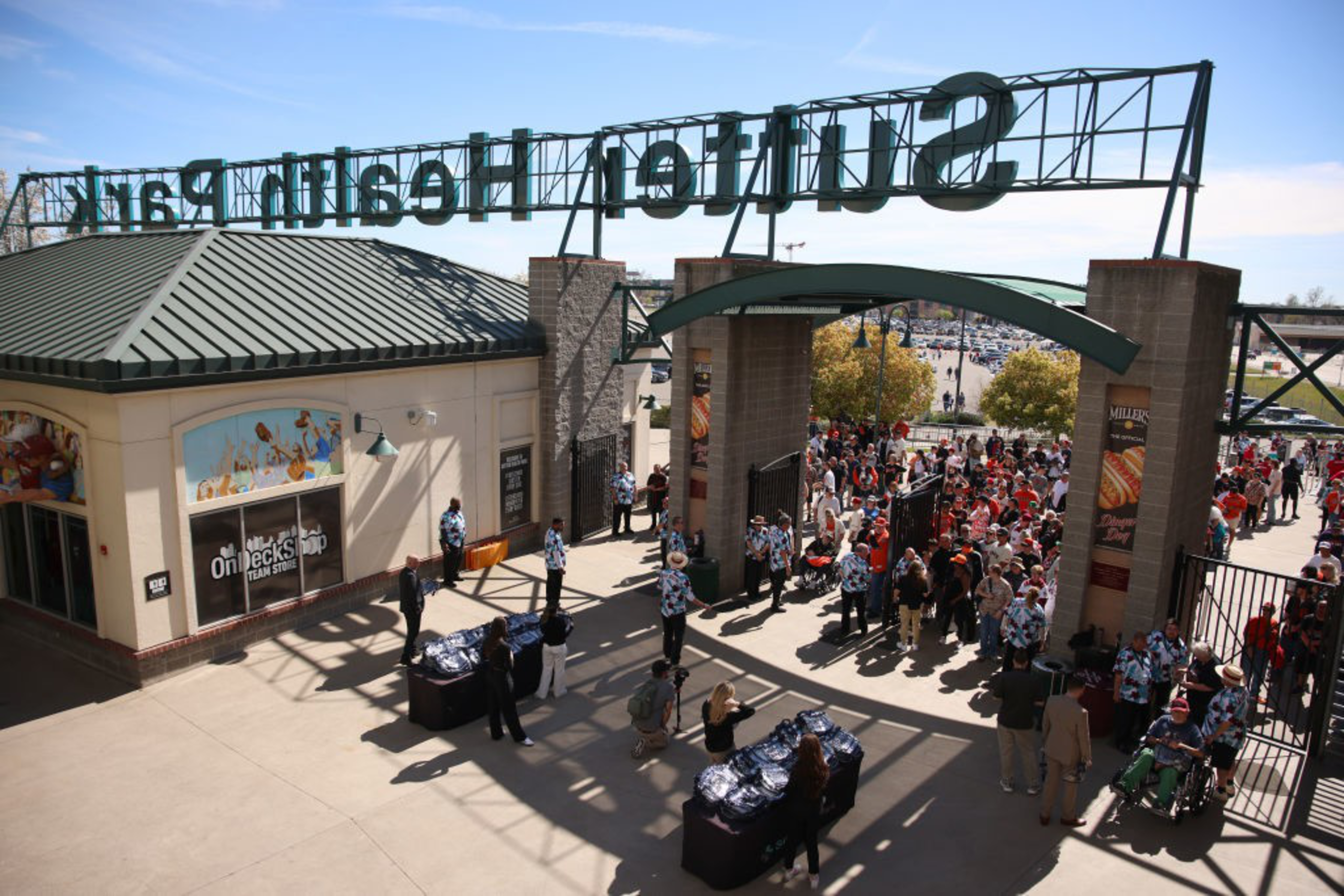 Crowds gather outside a ballpark entrance, with people in uniforms managing access. A team store is visible, and trees and a parking area are in the background.