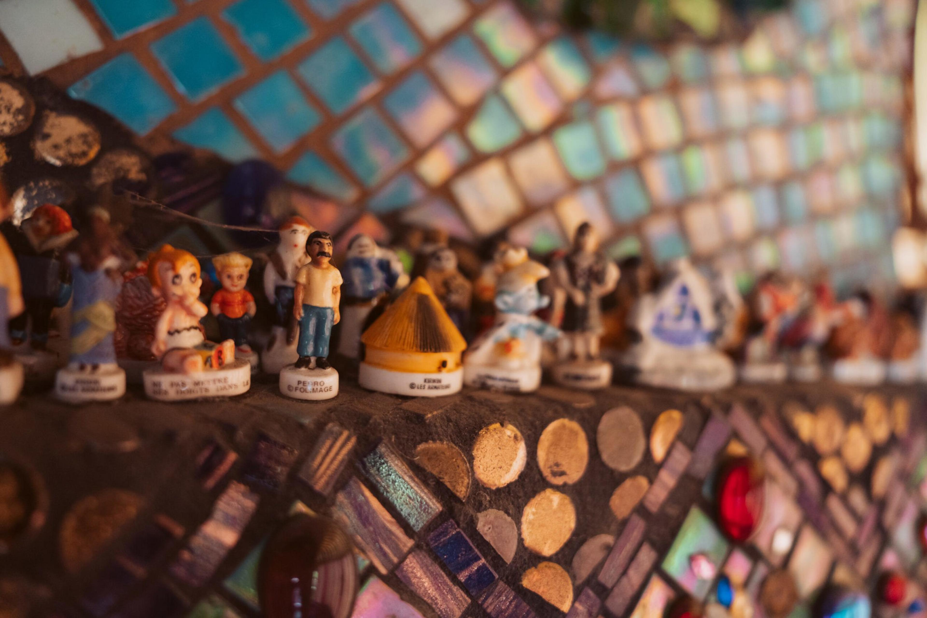 A row of small, colorful figurines stands on a mosaic shelf with blue, beige, and reddish-brown tiles creating a vibrant background.