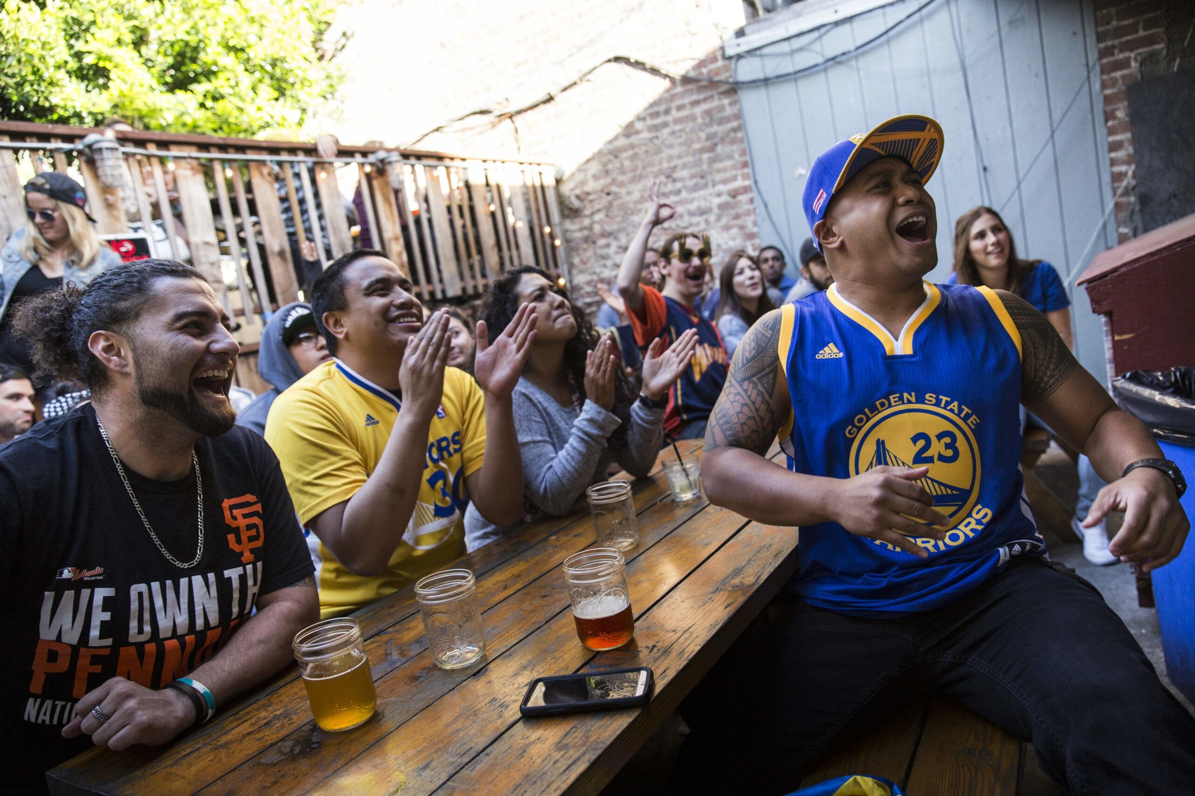 A diverse group of people cheer and laugh together at an outdoor wooden table, some wearing basketball jerseys and holding drinks.