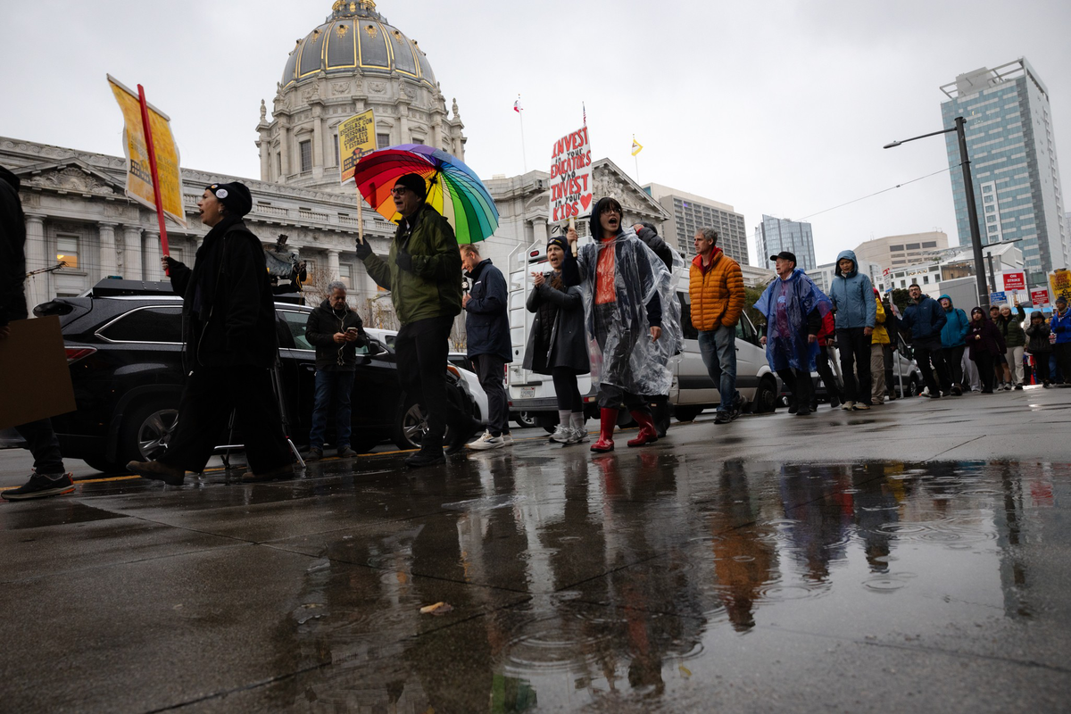 A group of people walk on a wet city sidewalk near a domed building, some wearing raincoats and holding signs, with reflections visible in puddles.