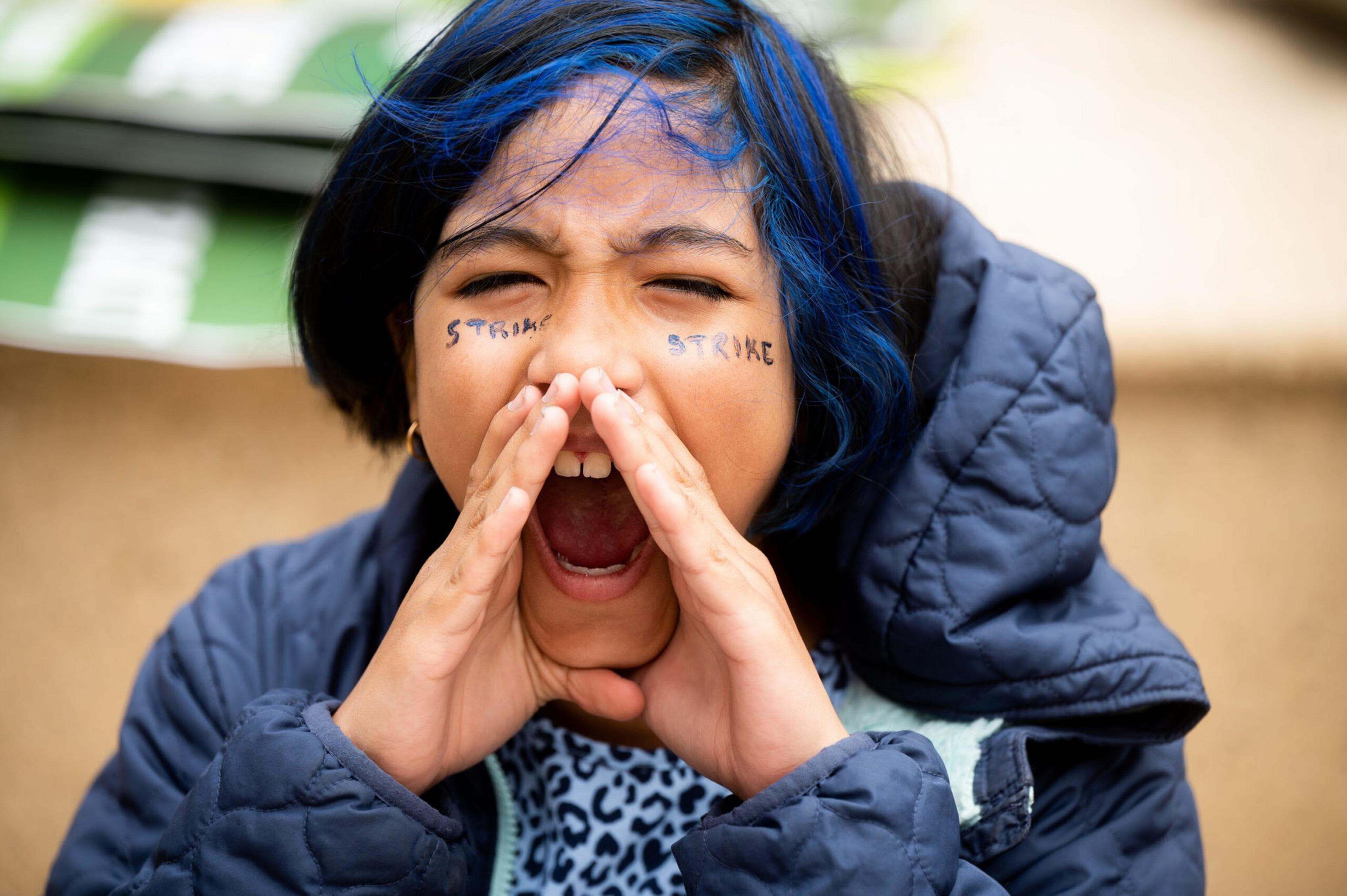 A person with blue hair and face paint saying "STROKE" on each cheek cups their hands around their mouth and shouts with eyes closed.