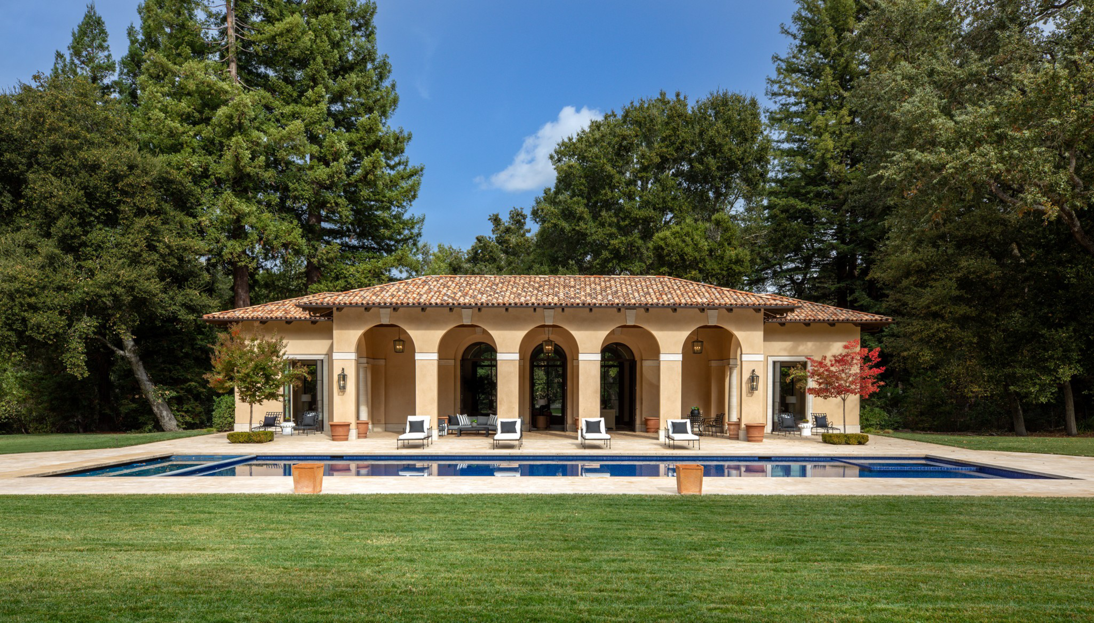 A single-story villa with a tiled roof features five arches facing a rectangular swimming pool, surrounded by a green lawn and tall trees.