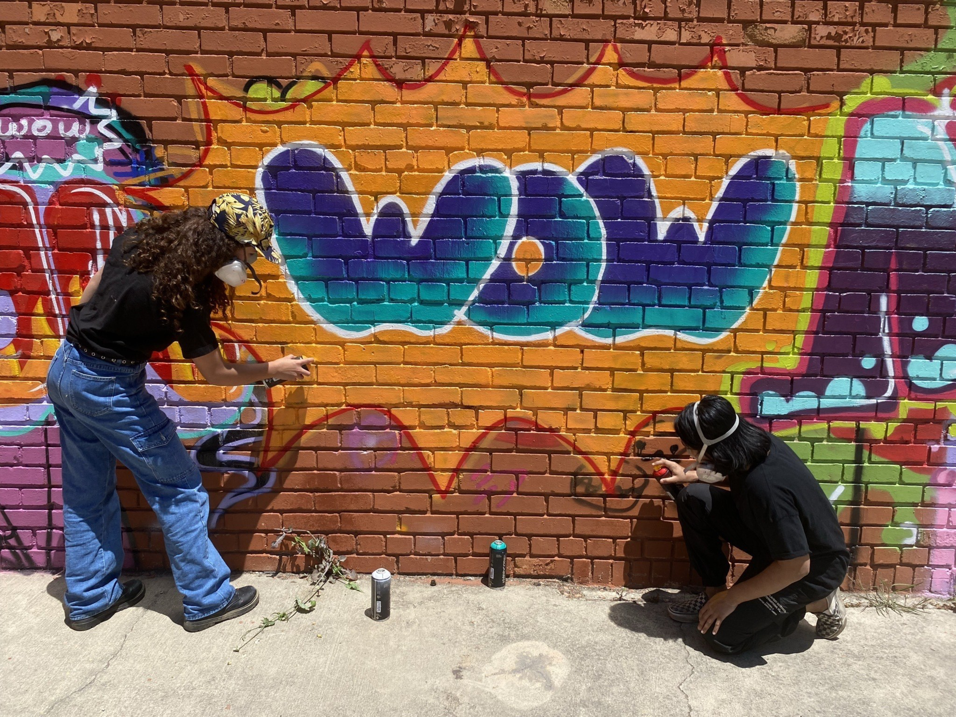 Two people wearing masks spray paint graffiti on a brick wall that says "WOW" in blue and teal letters with an orange burst background.