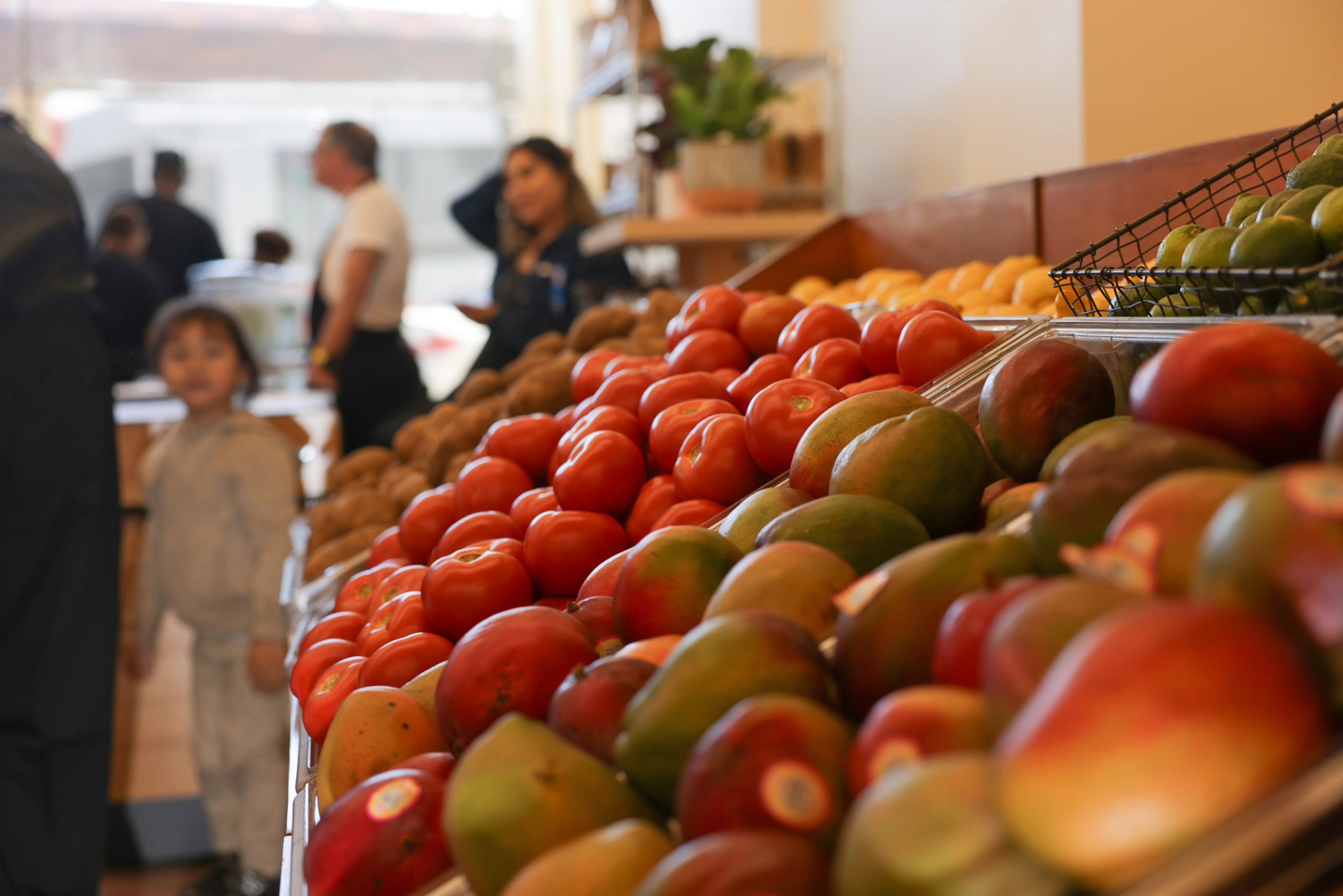 In a grocery store, colorful produce like tomatoes and mangoes are neatly displayed on a shelf, with people and a small child visible in the blurry background.