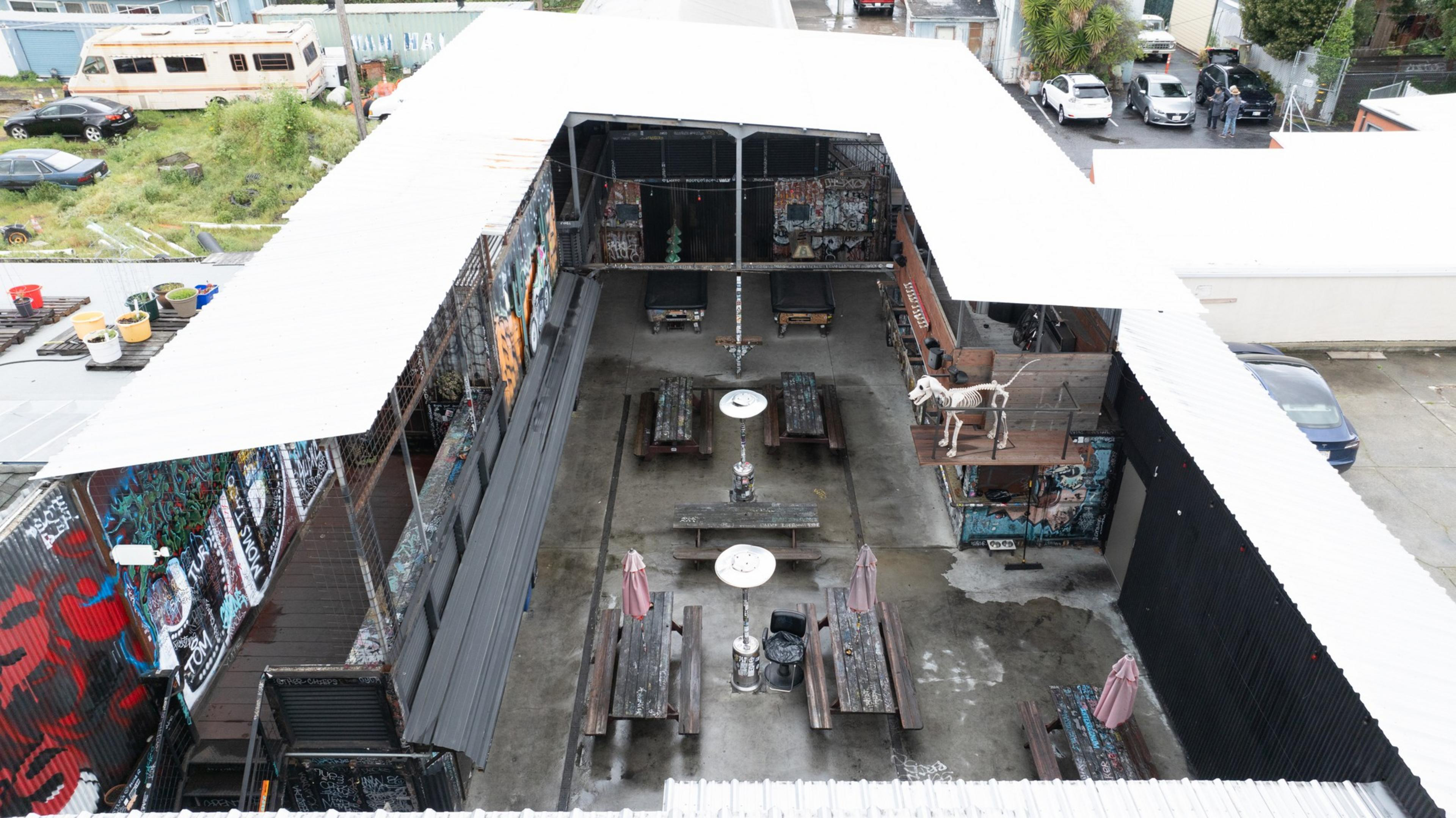 The image shows a U-shaped rooftop terrace with graffiti-covered walls and several picnic tables. A sculpture of a skeleton dog is on the right.