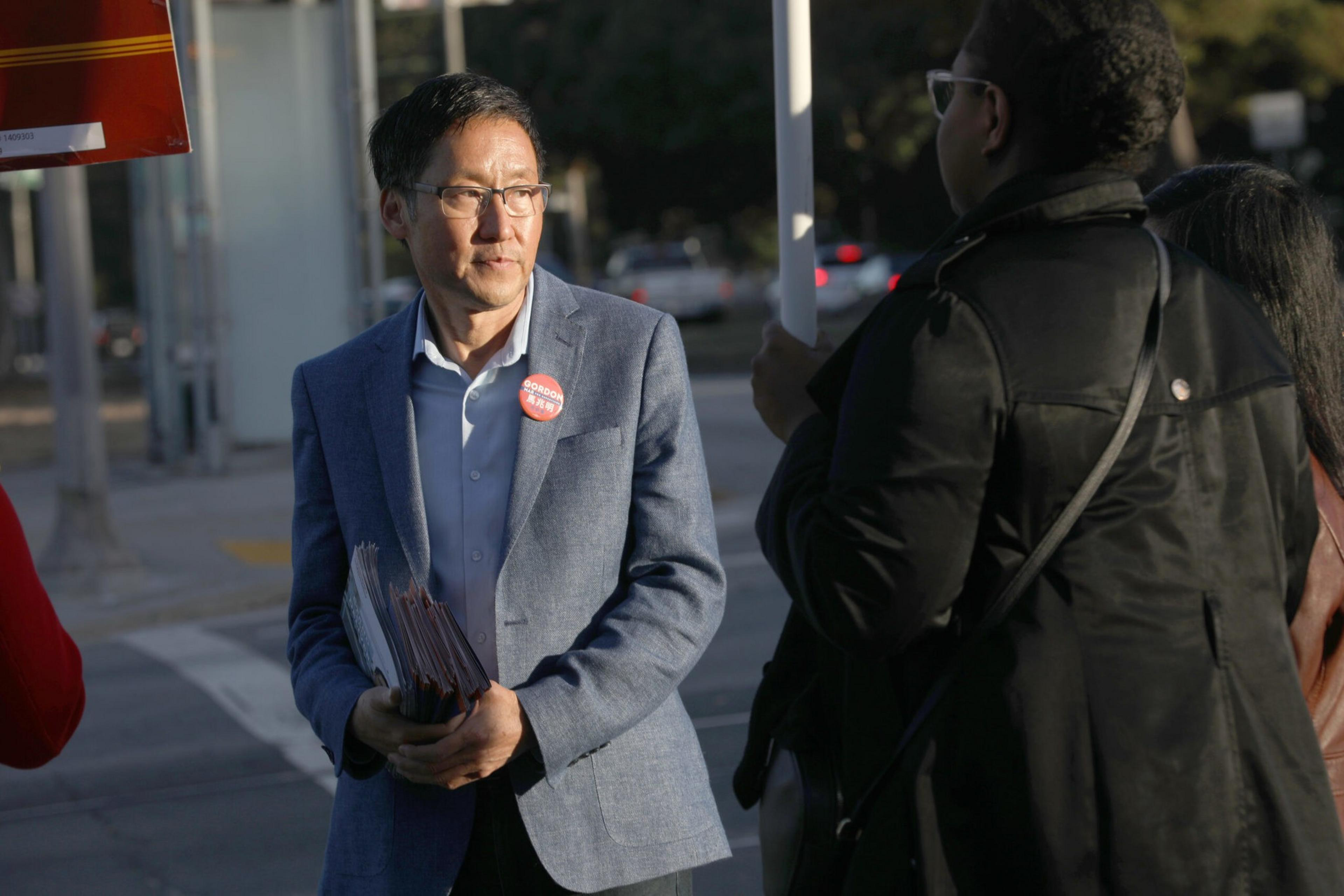 A man in a blue blazer and glasses holds papers and wears a red button, standing on a street corner with two people nearby.