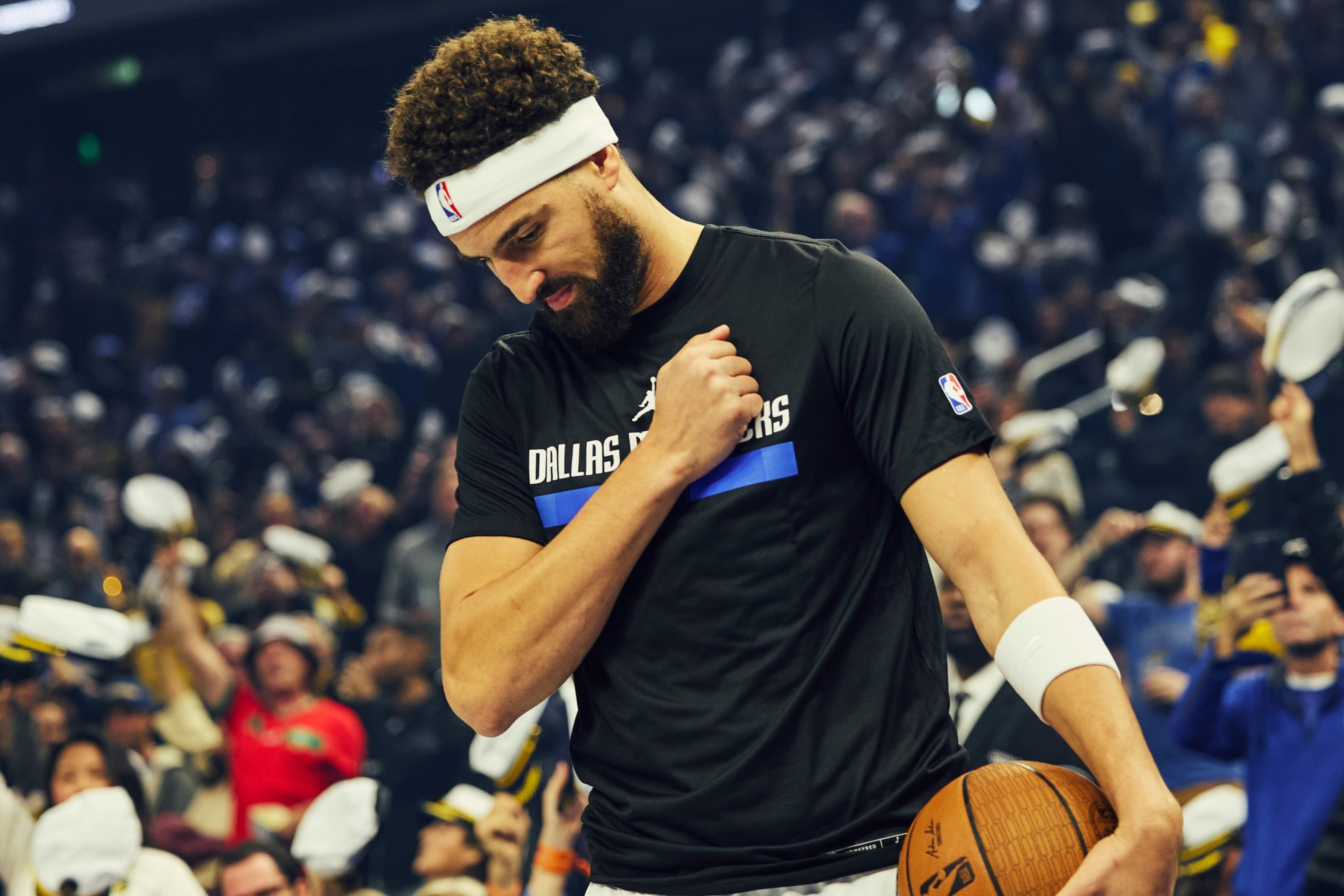 A basketball player wearing a white headband and a Dallas Mavericks shirt stands on a court, holding a ball, with a crowd in the background.