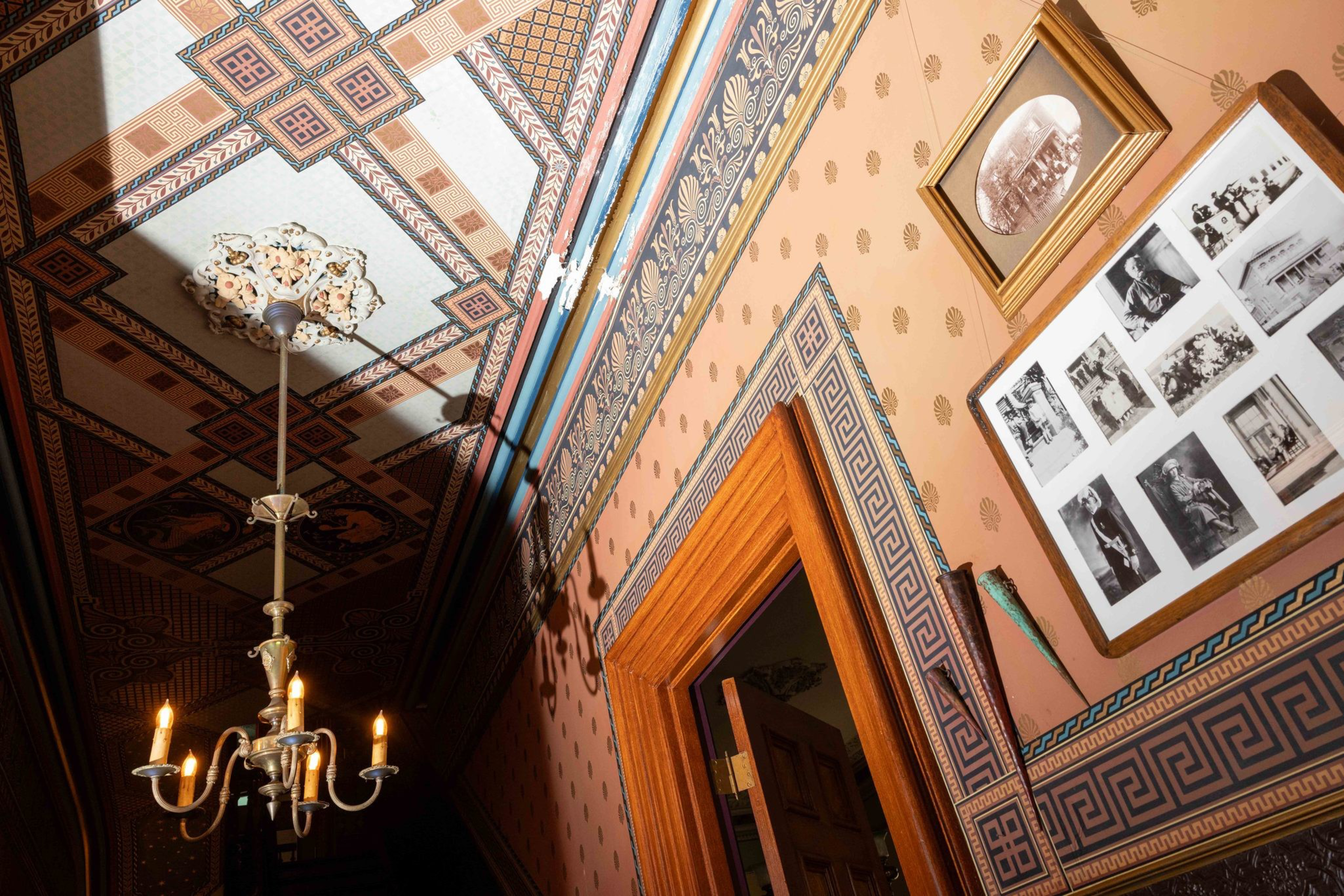 A warmly lit hallway features patterned walls and ceiling, a gold-framed photo collage, a single framed photo, and an antique chandelier with five candle-like bulbs.