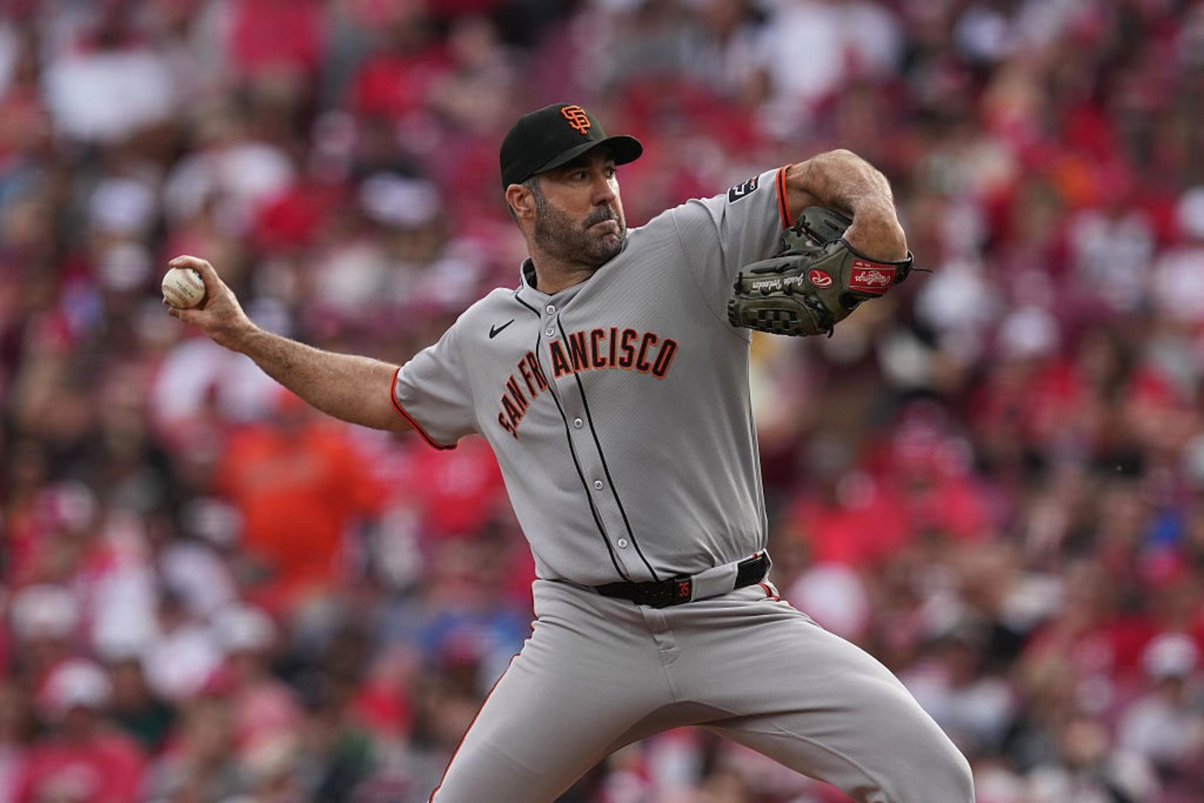A baseball pitcher in a gray "San Francisco" jersey is mid-throw, holding a ball in his right hand. The crowd is a blur of red and white in the background.
