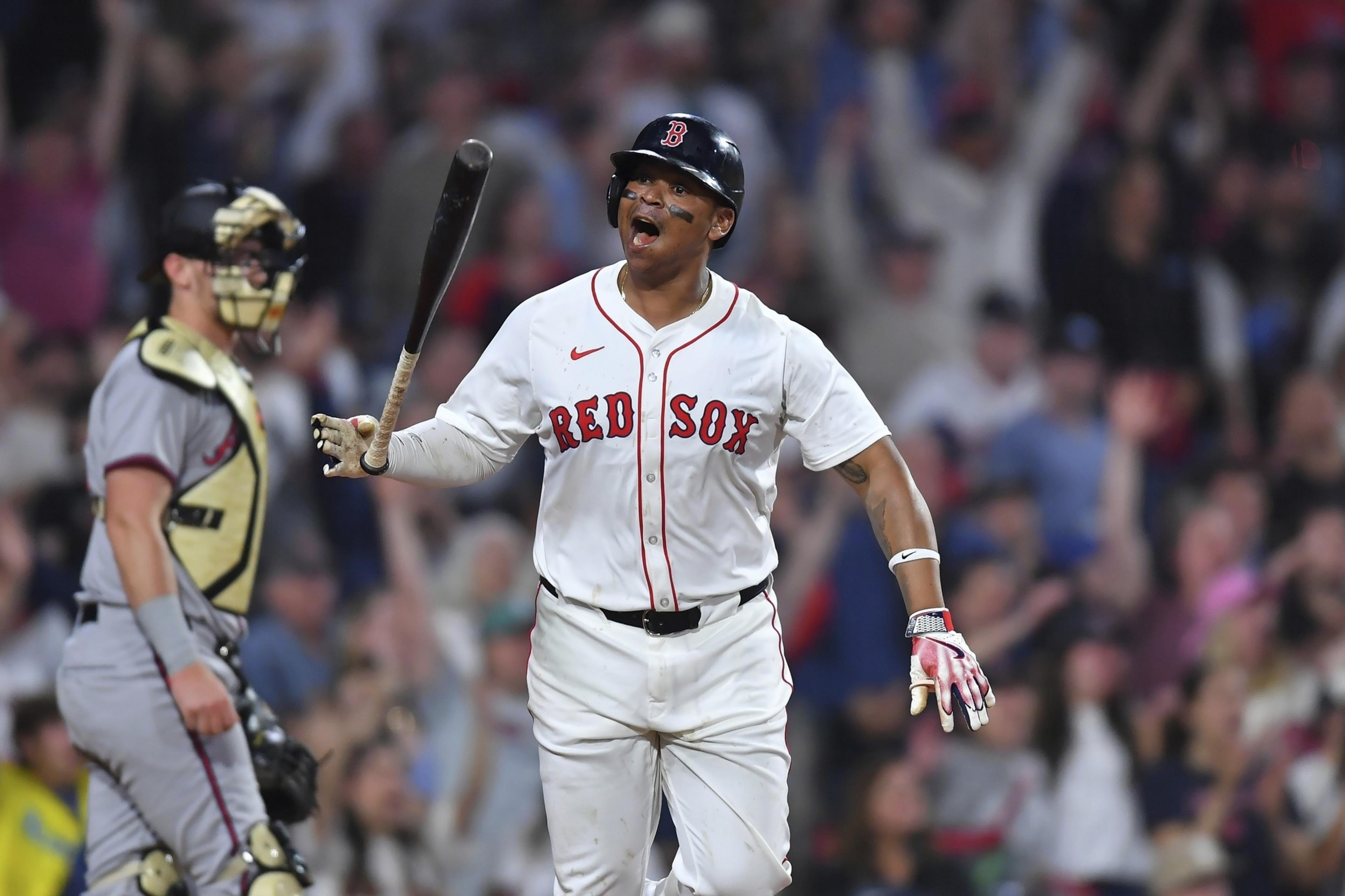 A baseball player in a Red Sox uniform stands excitedly on the field, holding a bat. A catcher is nearby, and the crowd is visible in the background.