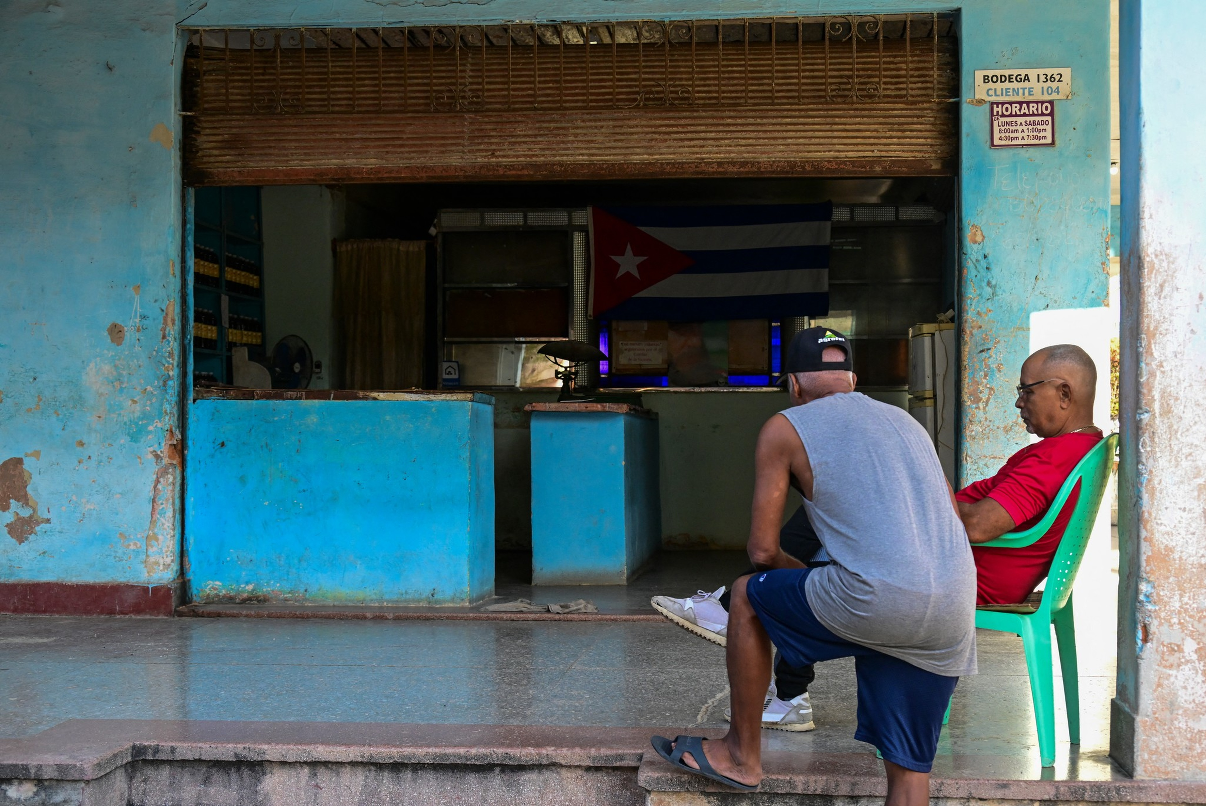 Two men sit outside a worn blue storefront with a Cuban flag hanging inside, one on a green plastic chair, the other sitting on the step.