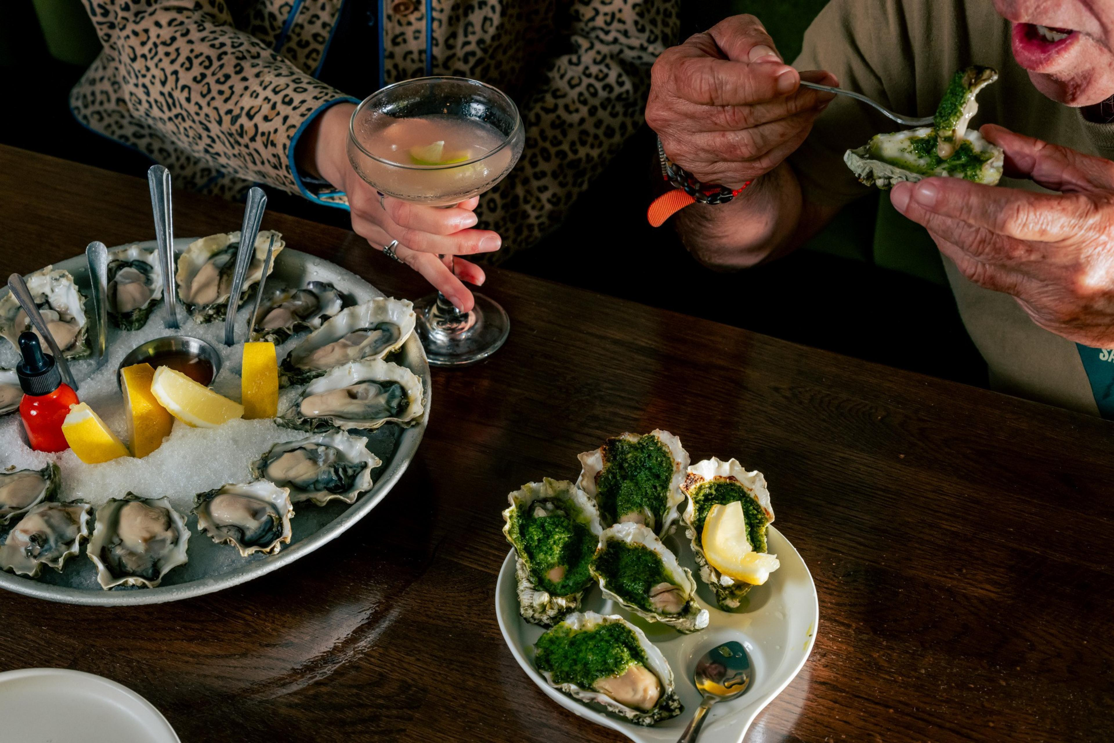 A person is about to eat oysters, surrounded by two plates of them. One plate has classic oysters with lemon and sauce; the other plate has oysters topped with a green garnish. Another person holds a martini.