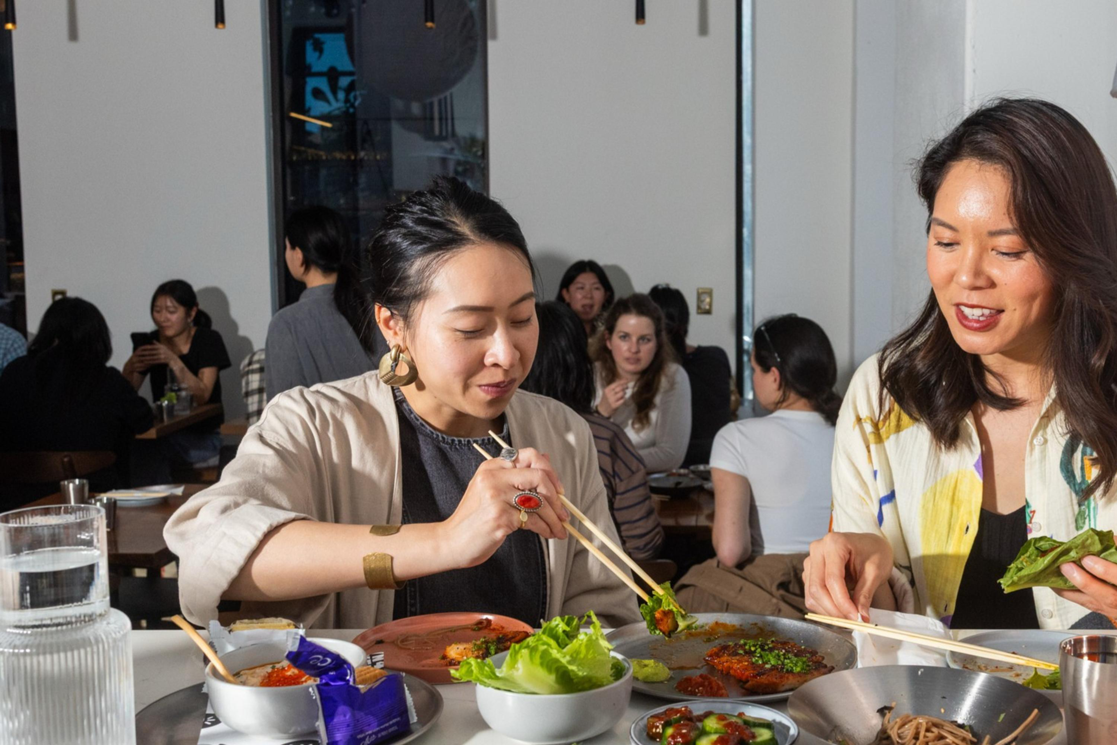 Two women sit at a table enjoying a meal with various dishes. They're using chopsticks, surrounded by plates of food, a candle, and a bright orange lamp.