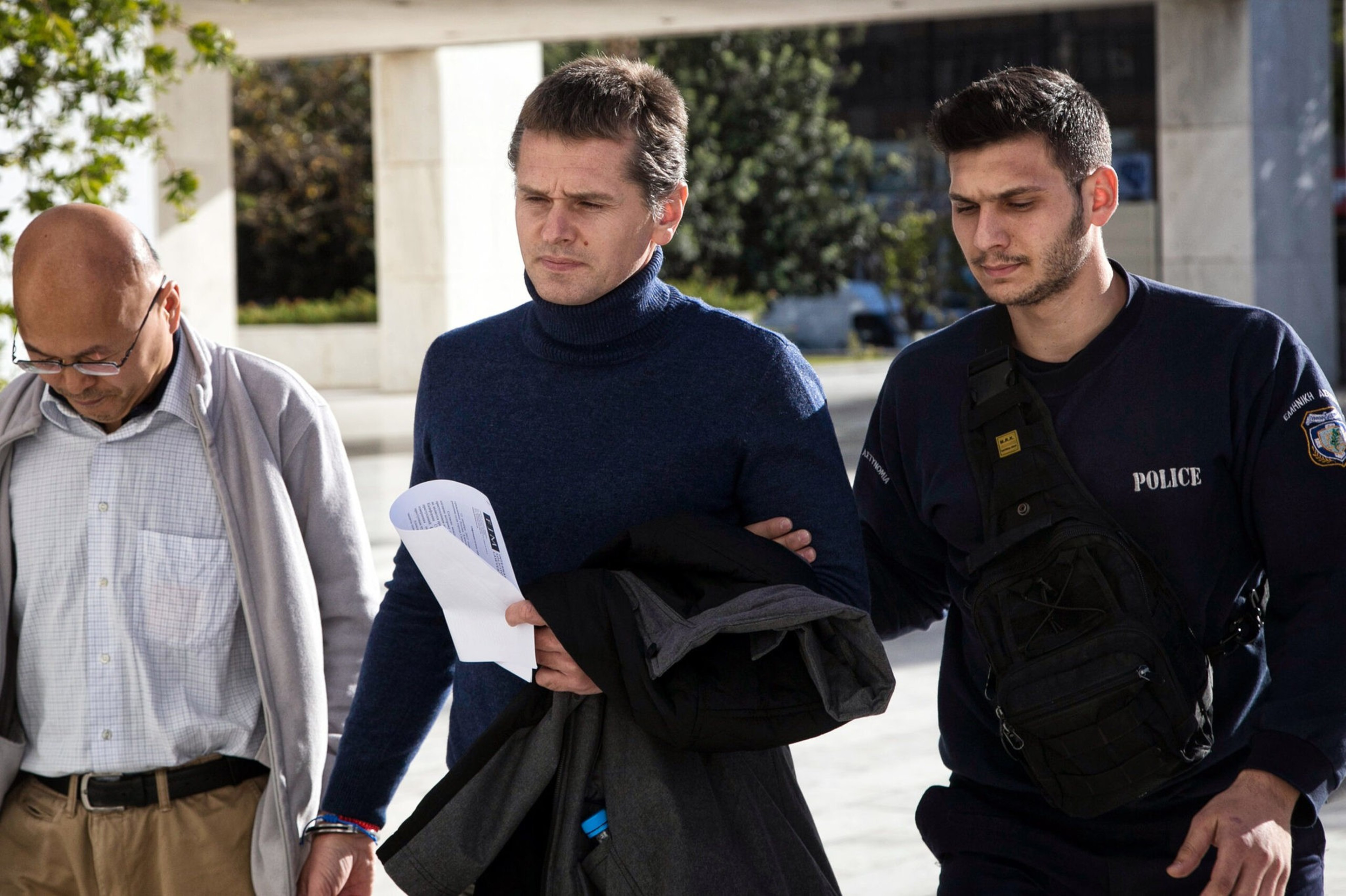 A man in a blue sweater is being escorted by a police officer and another man outdoors, holding a paper and a jacket.