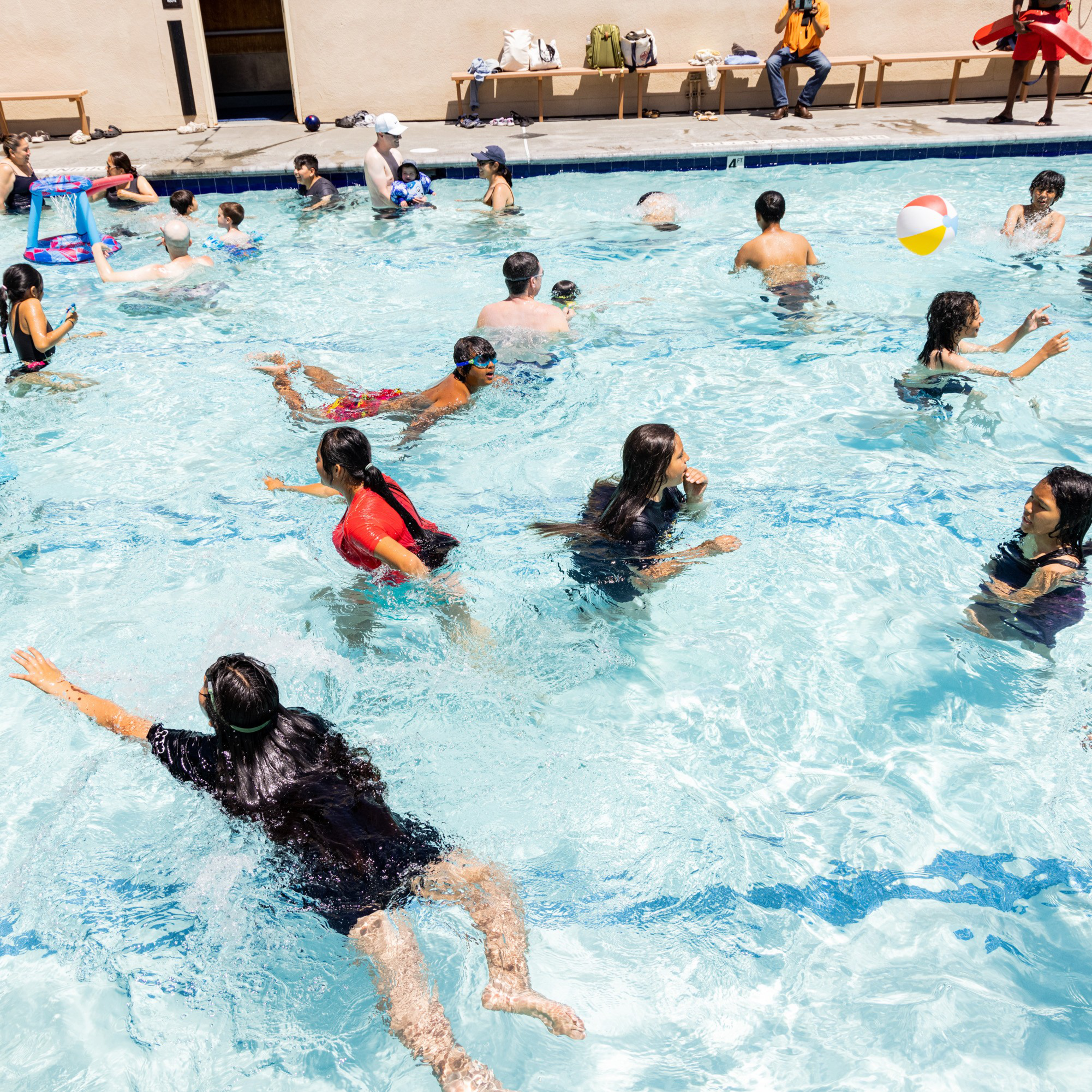 A busy pool scene with numerous people swimming and playing, including some children. A colorful beach ball is in the air, and a few people are seated on benches outside the pool.