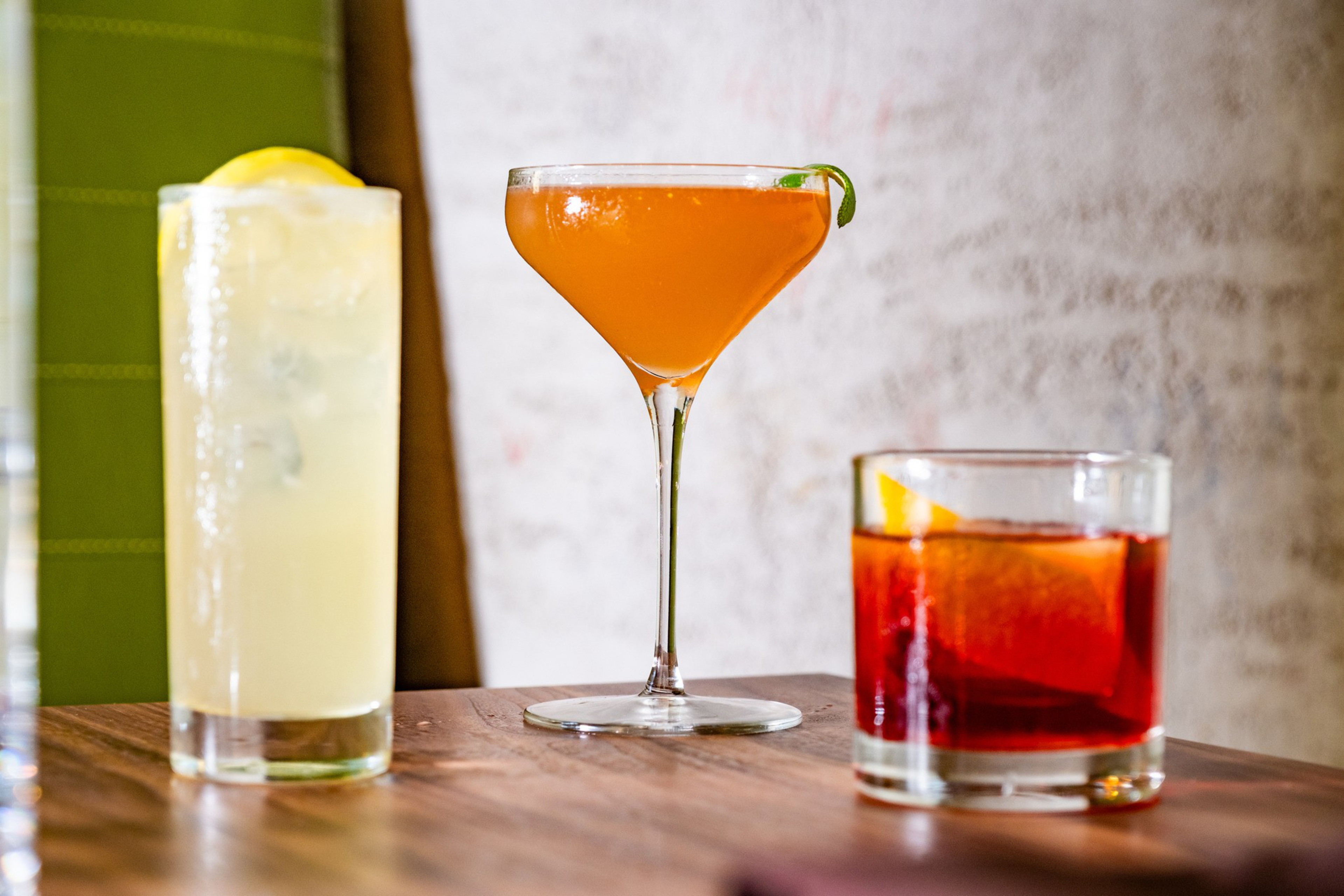 Three drinks are on a table: a tall glass with a lemon slice, a martini glass with an orange drink and lime garnish, and a rocks glass with an orange slice.