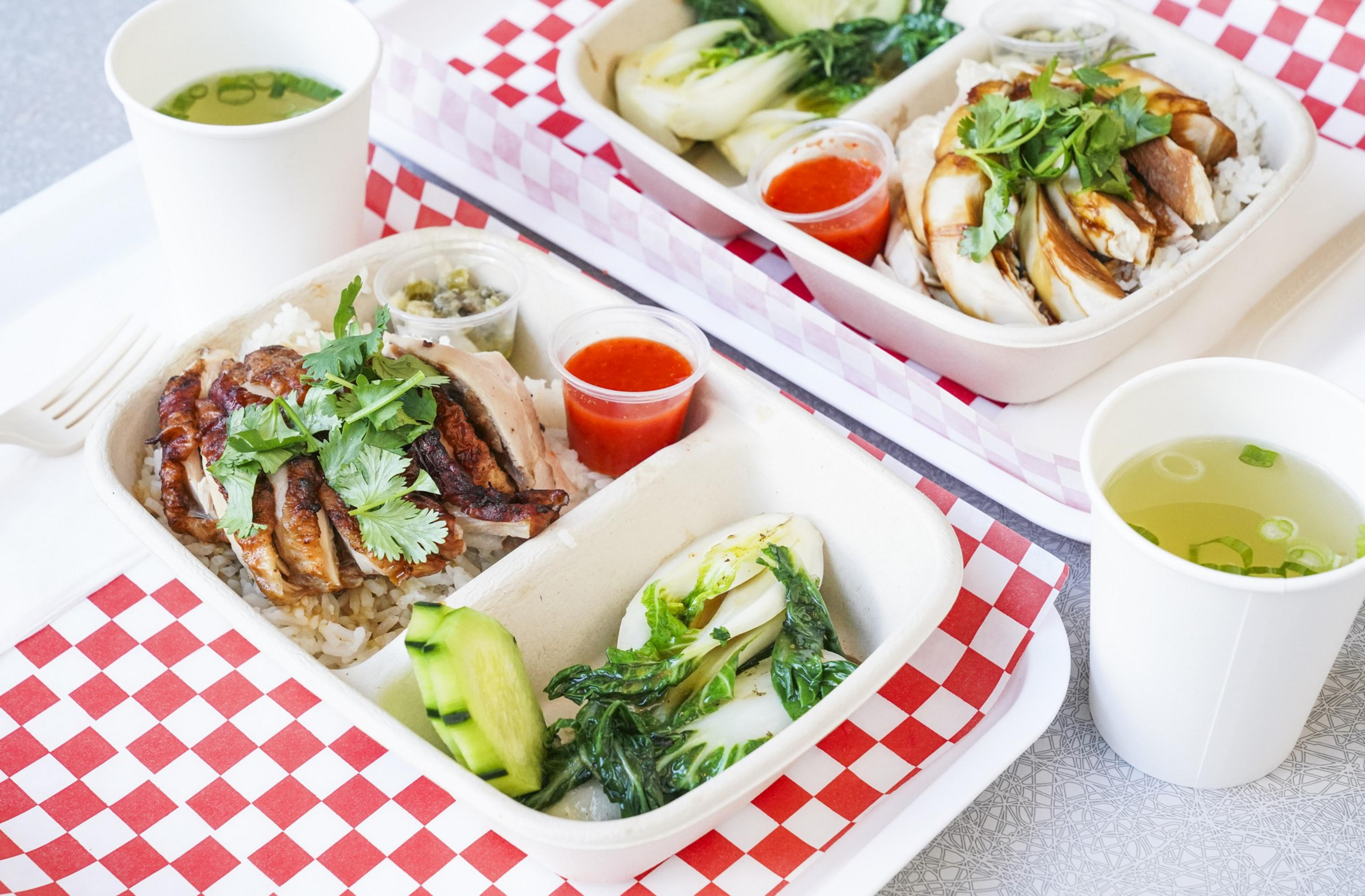Two trays contain rice, grilled chicken with cilantro, vegetables, sauce cups, and a small soup cup. The setup is on red checkered paper.
