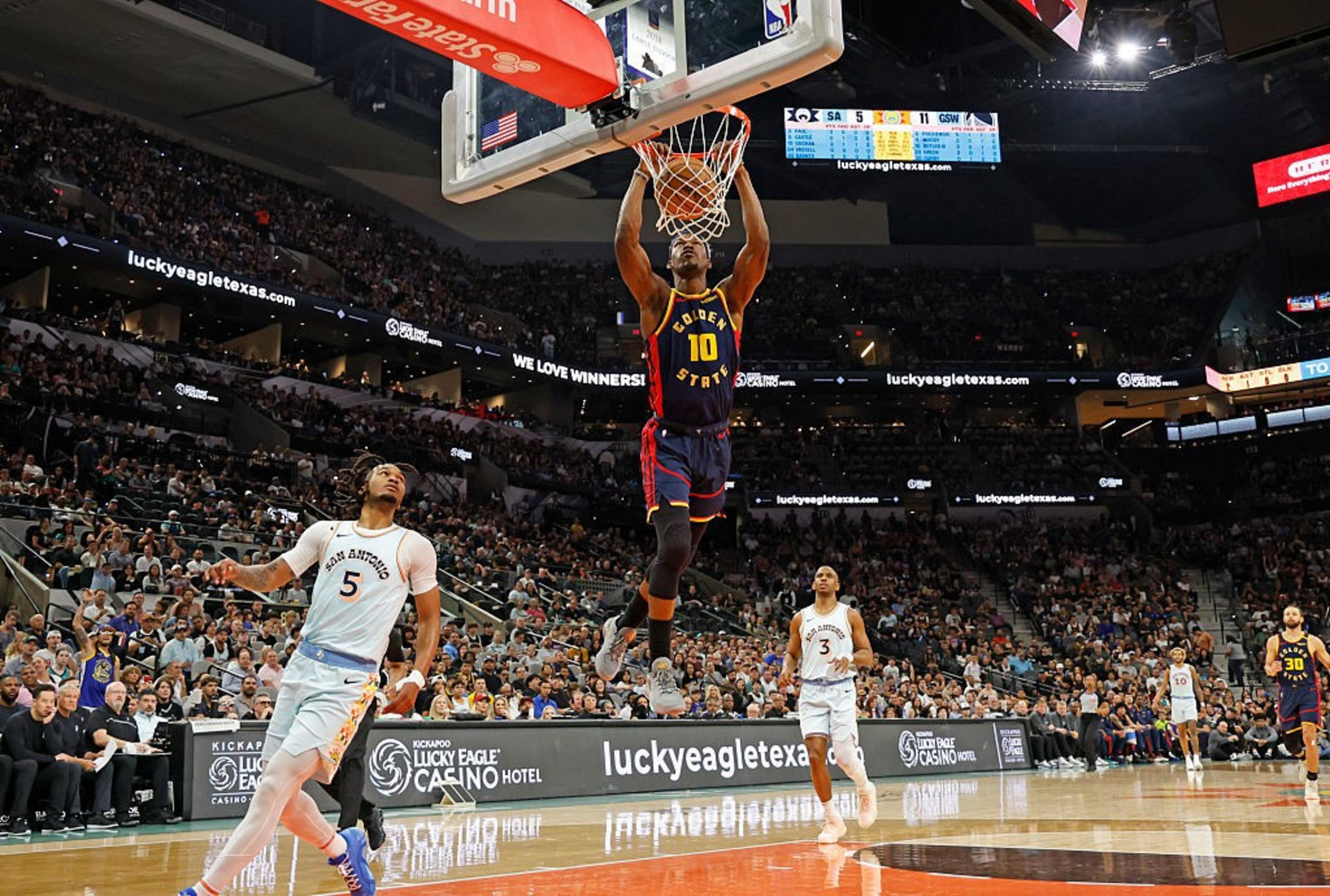 A basketball player in a Golden State uniform is dunking the ball, with opposing San Antonio players watching. The arena is crowded with spectators.
