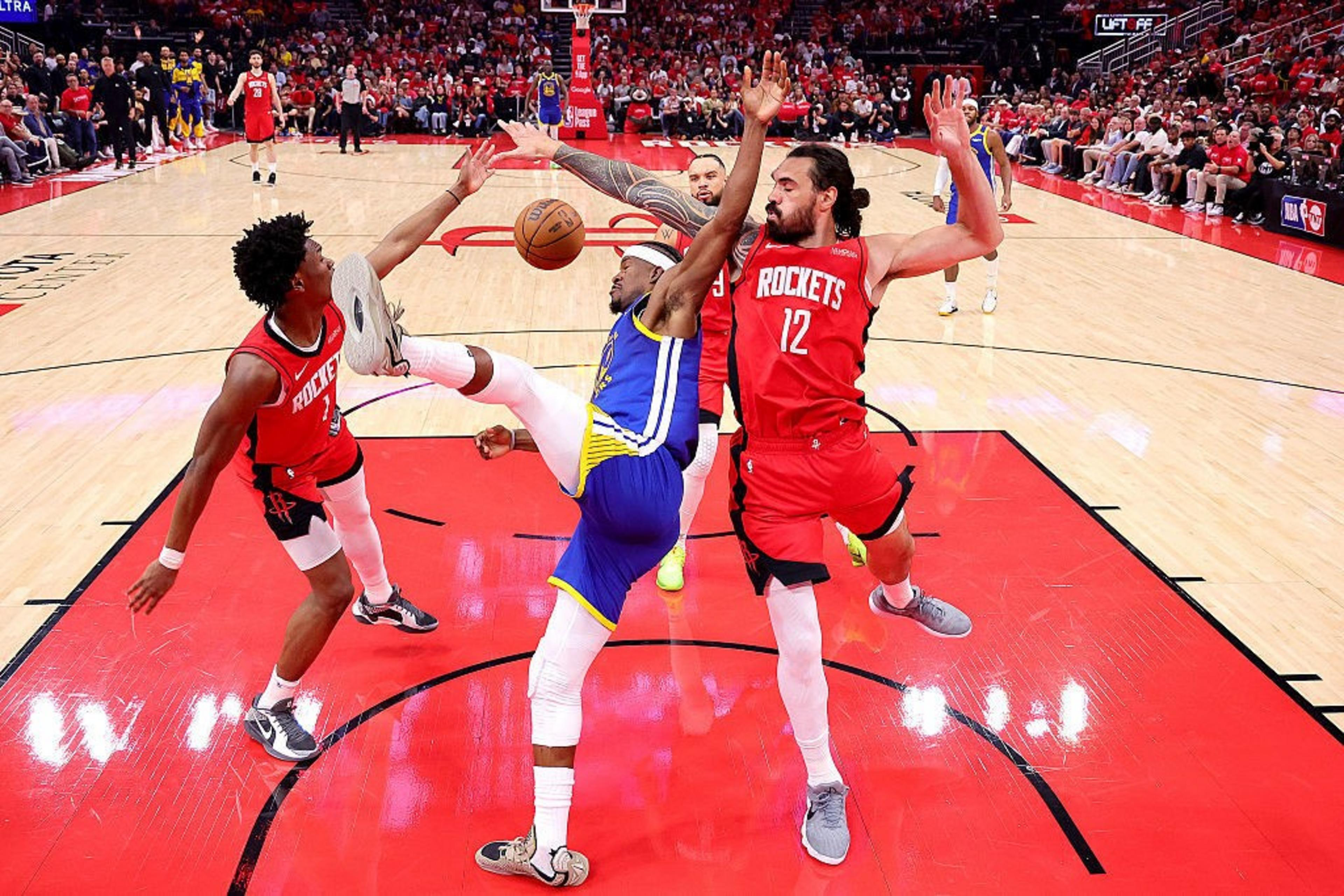 A basketball player in a blue uniform attempts a shot while dodging two players in red uniforms. The scene is tense, with arms and legs entangled.