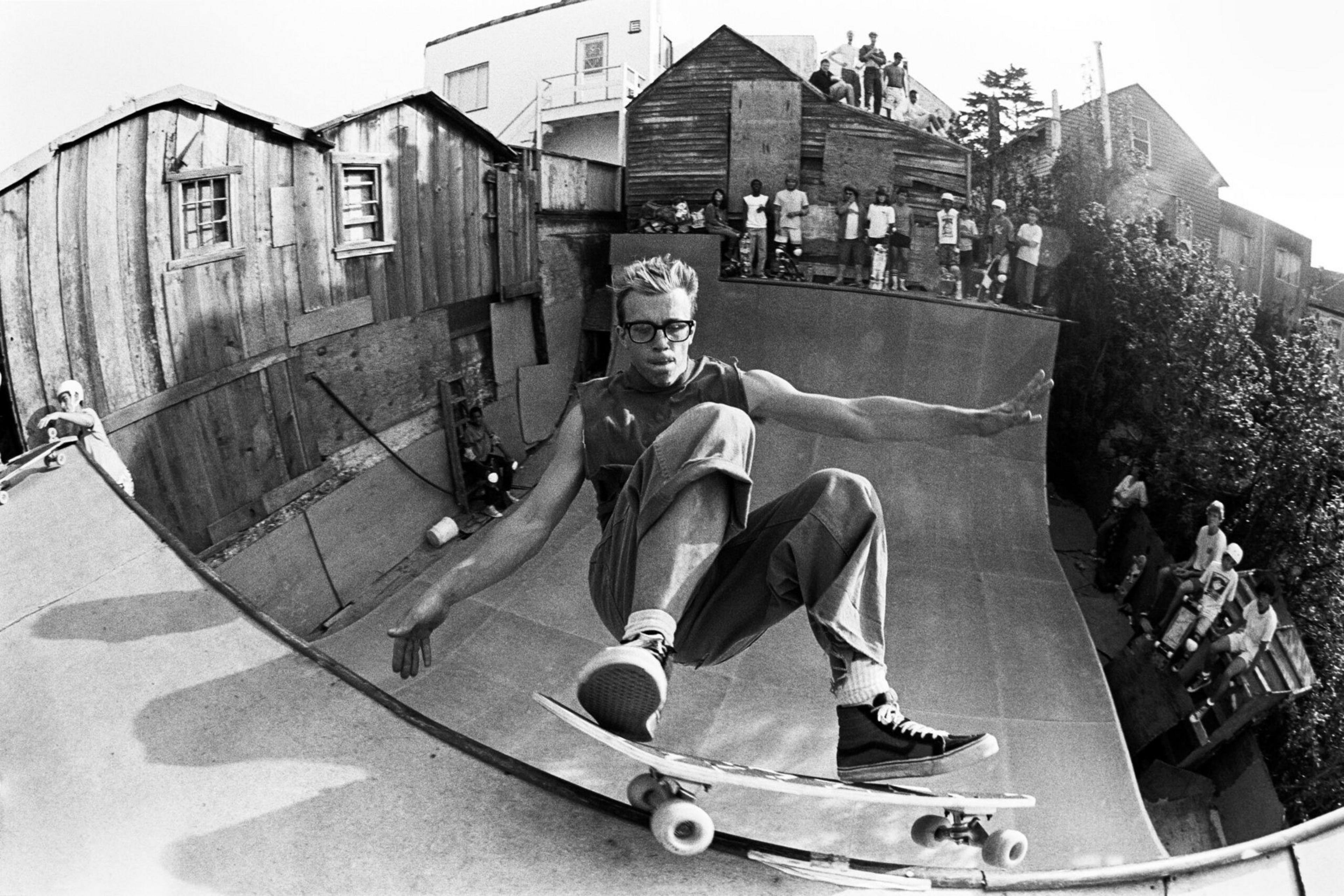 A skateboarder on a halfpipe with onlookers above; old wooden shacks nearby. Black and white photo.
