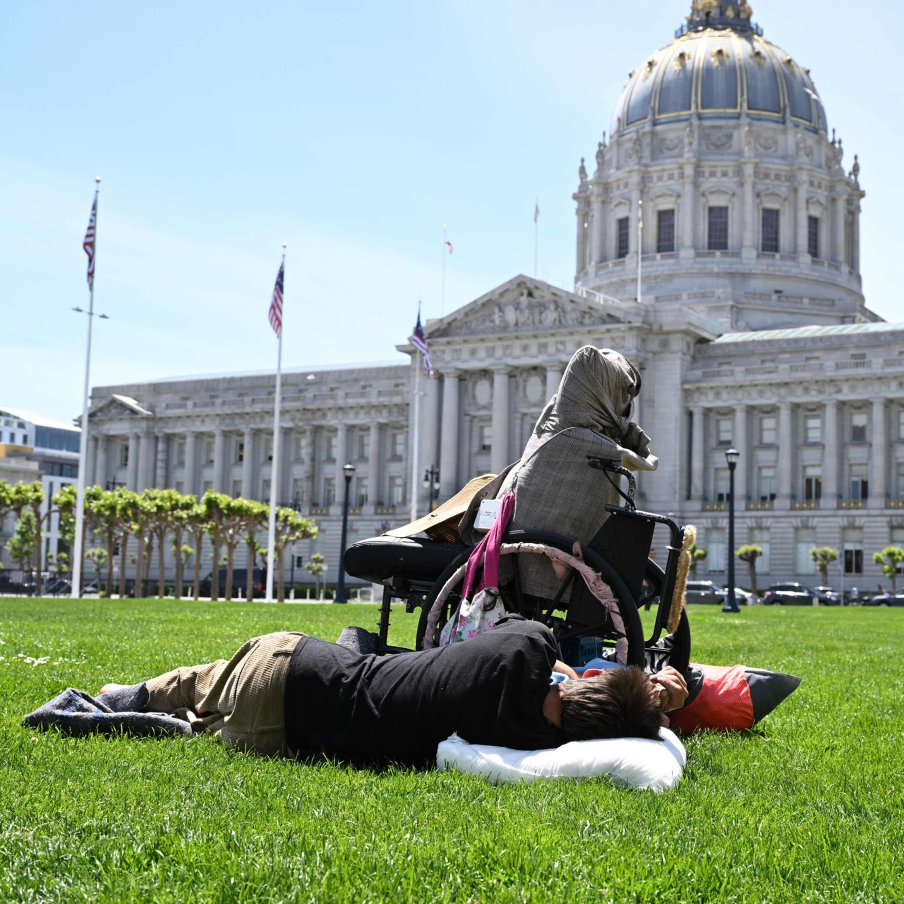 A person lies on the grass using a pillow near a wheelchair, with a large classical building featuring a dome in the background.