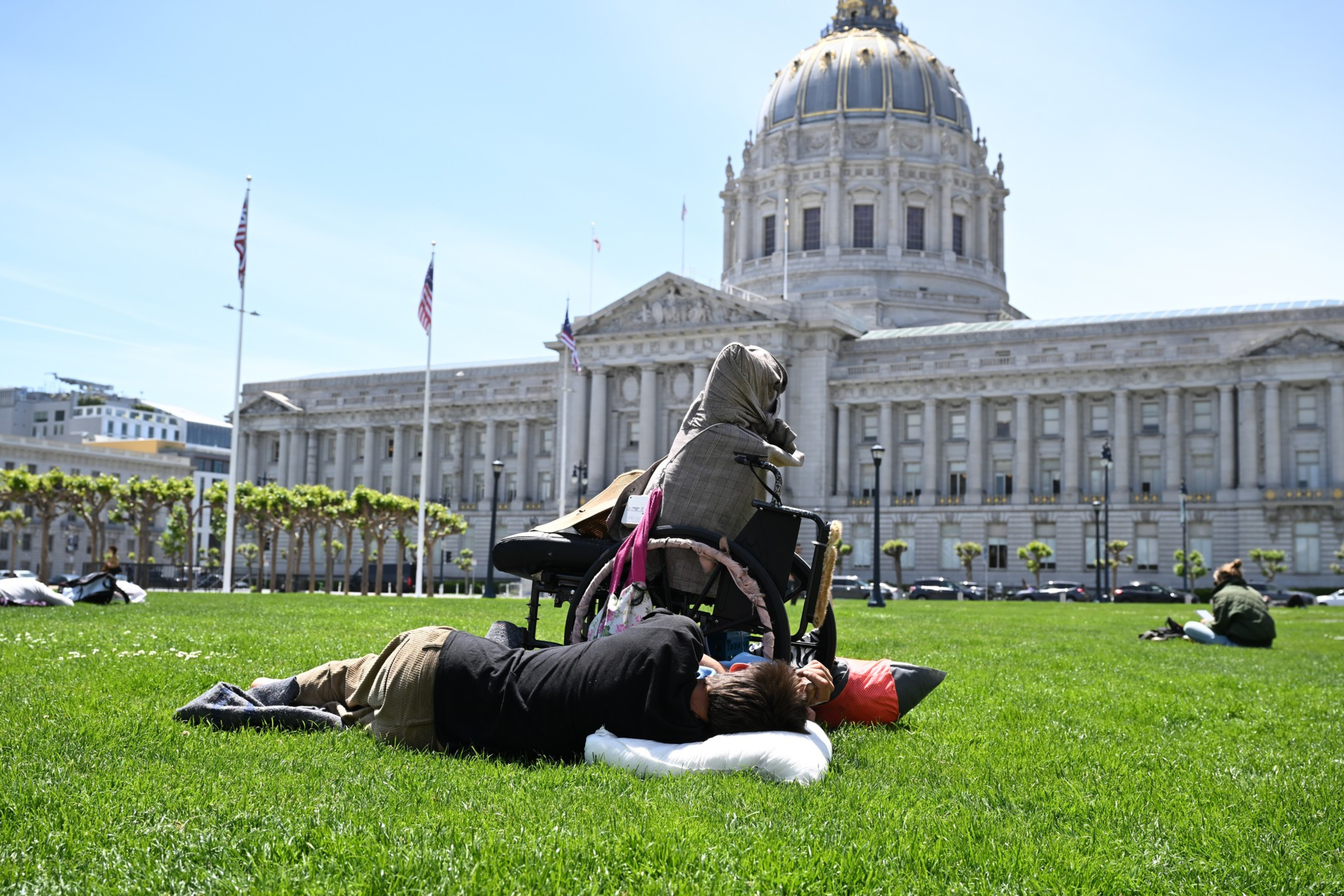 A person lies on the grass using a pillow near a wheelchair, with a large classical building featuring a dome in the background.