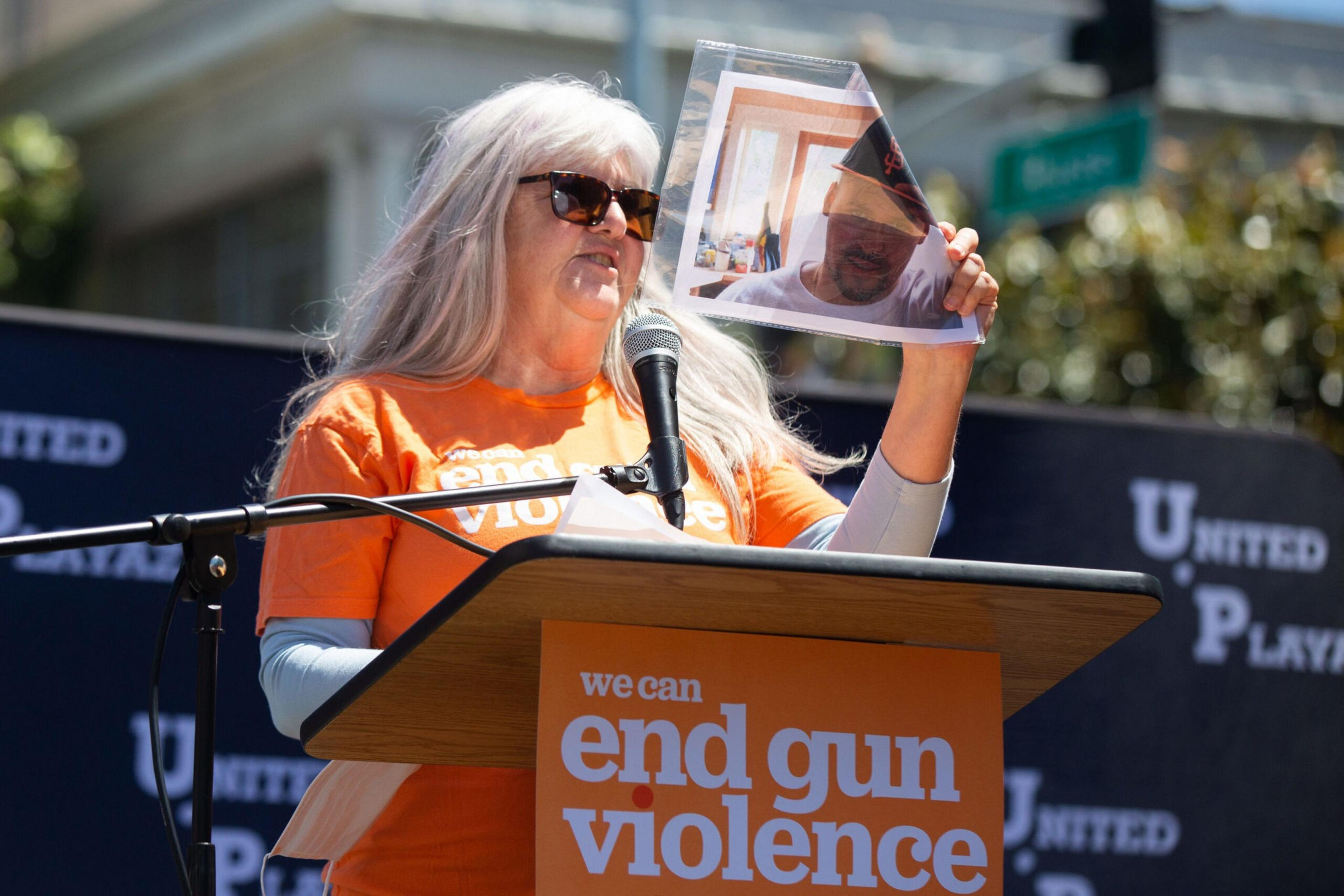 A woman in an orange "end gun violence" shirt and sunglasses speaks at a podium while holding a photo of a man wearing a baseball cap.