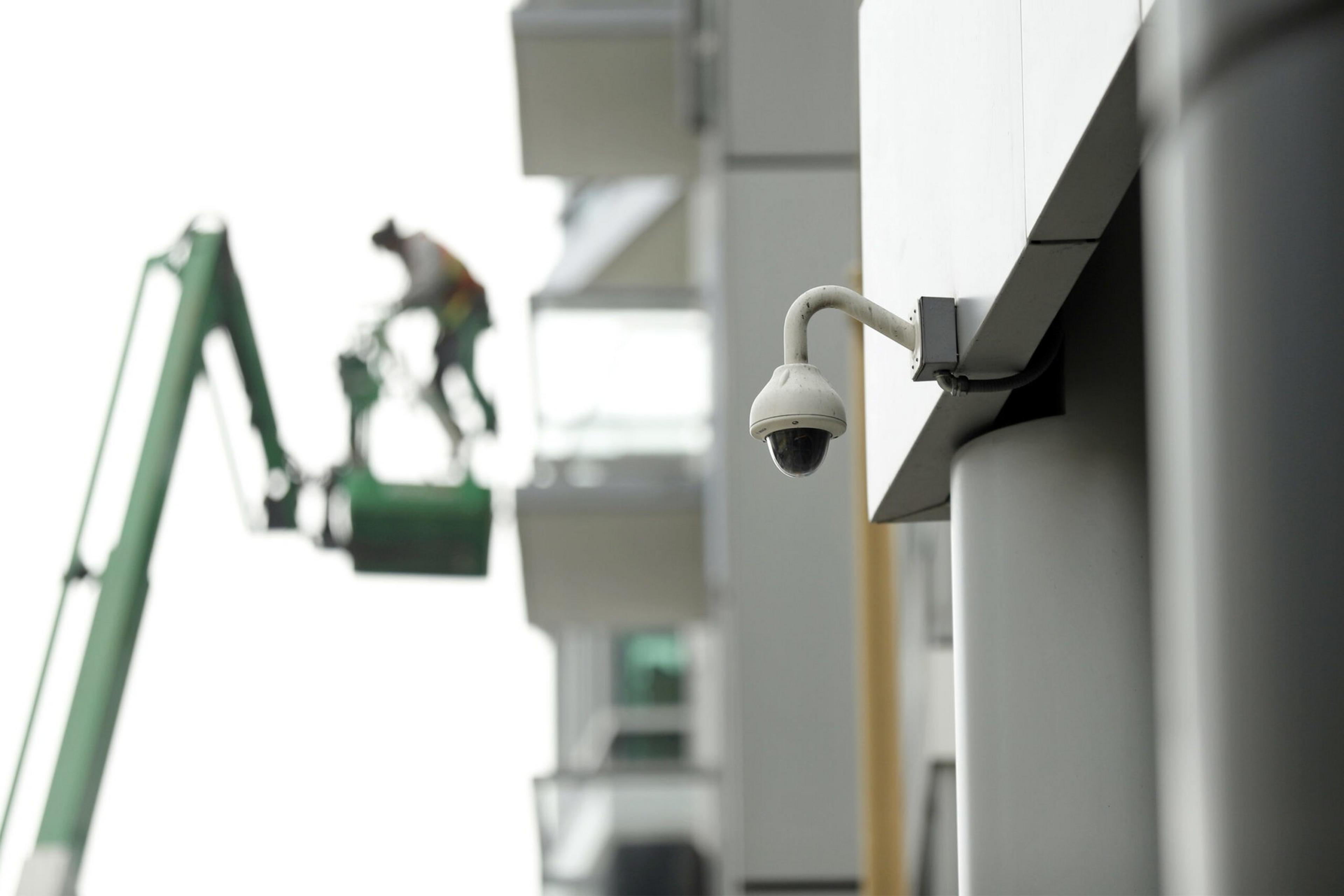A security camera mounted on a building focuses on a worker in safety gear standing on a green elevated platform near a multi-story building.