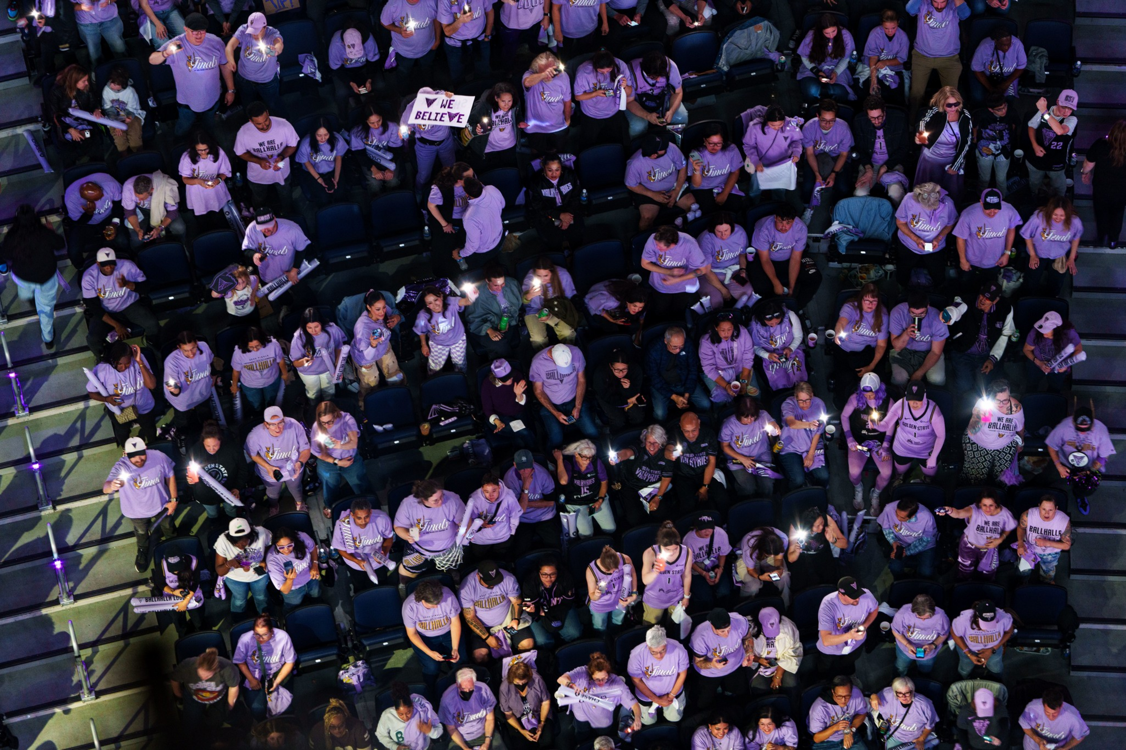 A large crowd seated in an arena, mostly wearing purple shirts, some holding lights and a sign that says "WE BELIEVE."
