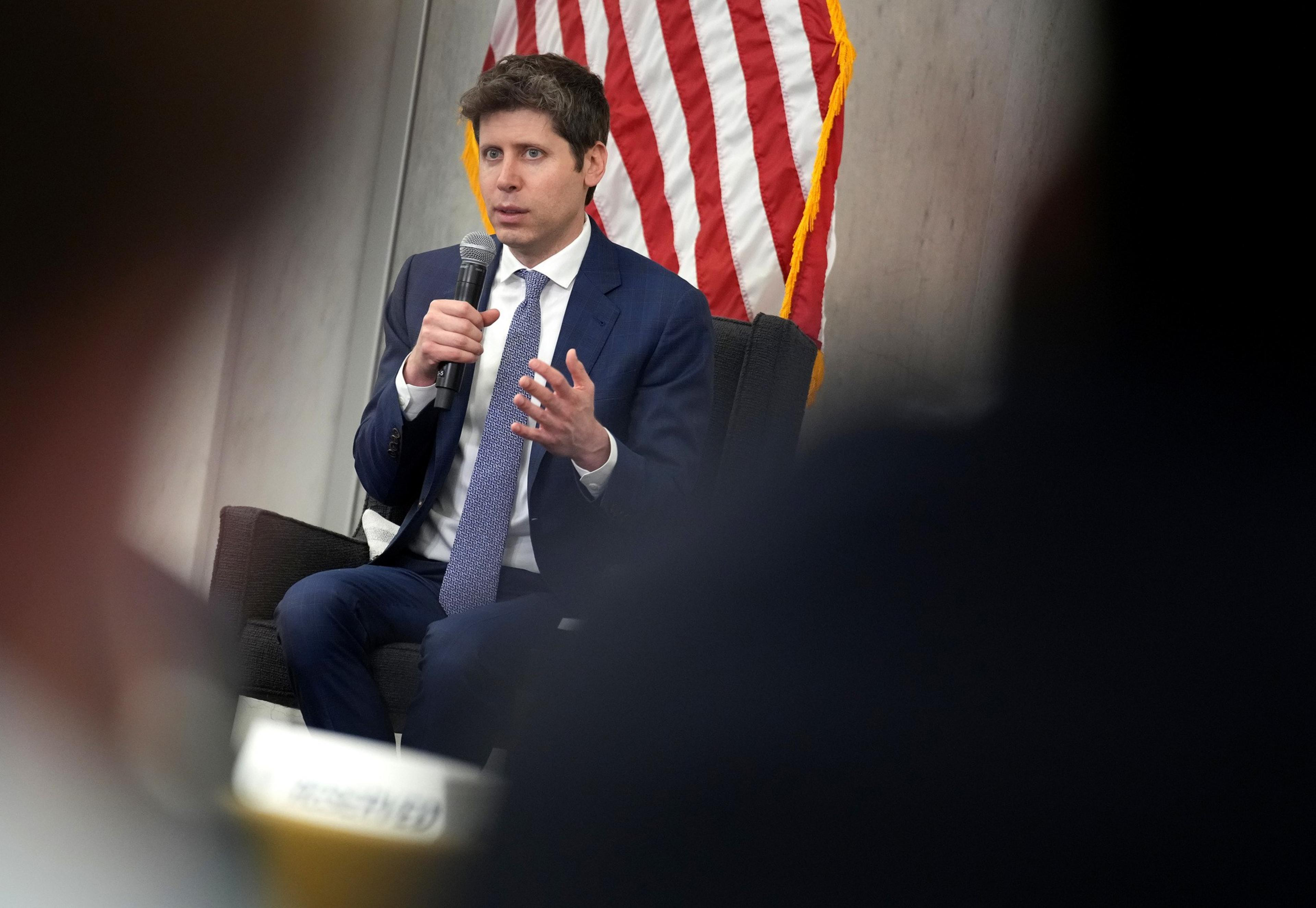 A man in a blue suit and tie holds a microphone and speaks, seated in front of a US flag with blurred audience members in the foreground.