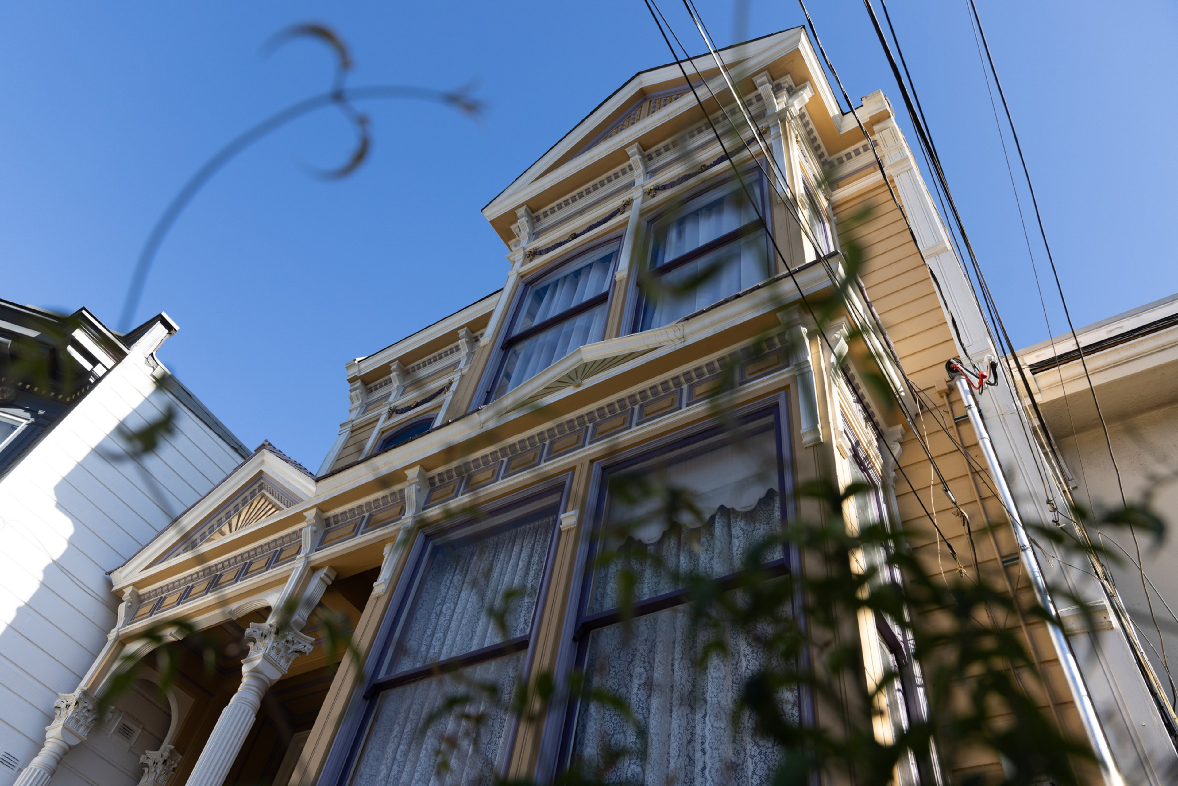 The image shows a detailed, ornate Victorian house against a clear blue sky, with decorative trim, large windows with lace curtains, and some foliage in the foreground.