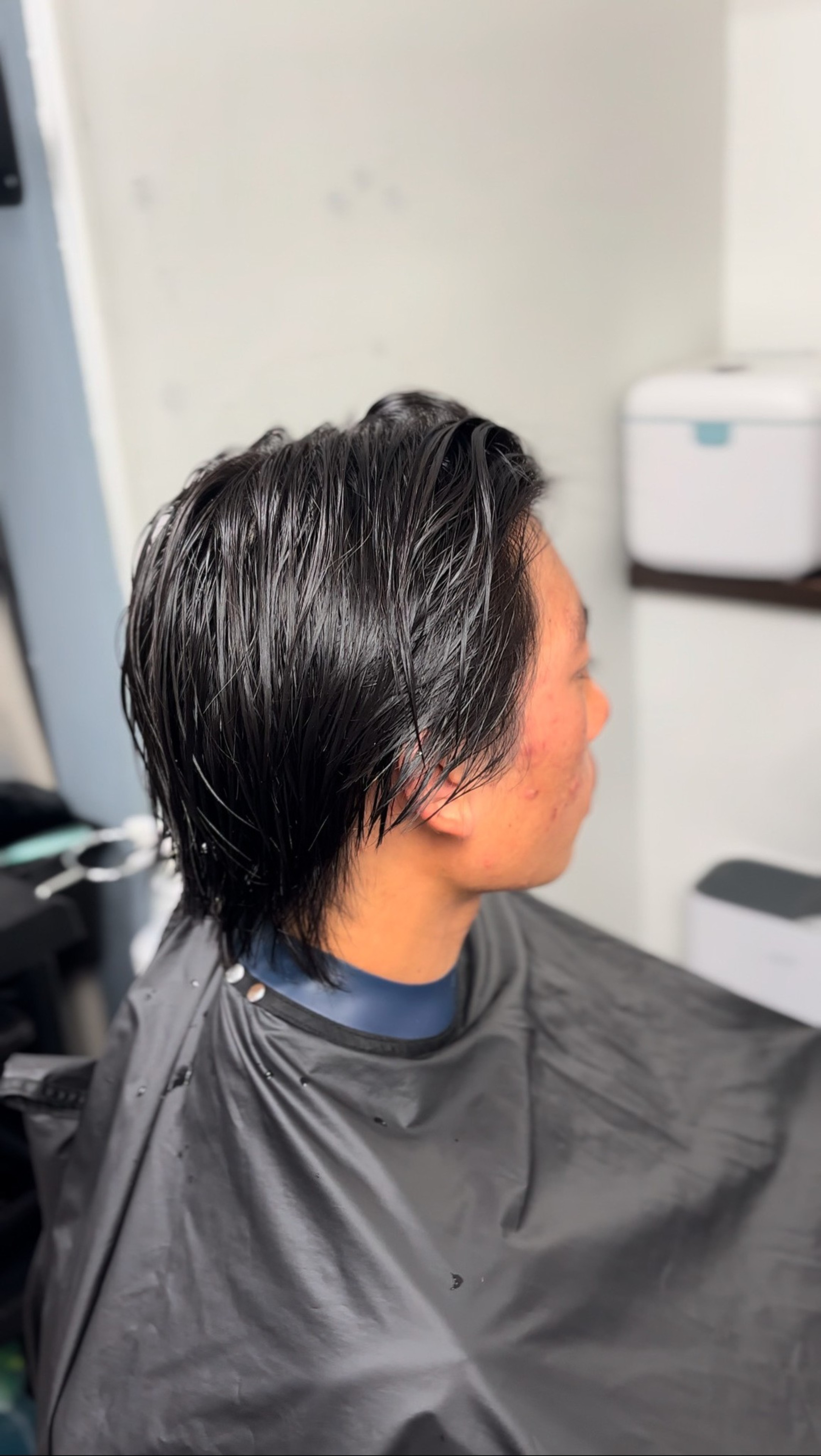 A person with wet, dark hair sits in a salon chair, wearing a black cape. The background is simple and contains a shelf and a small appliance.