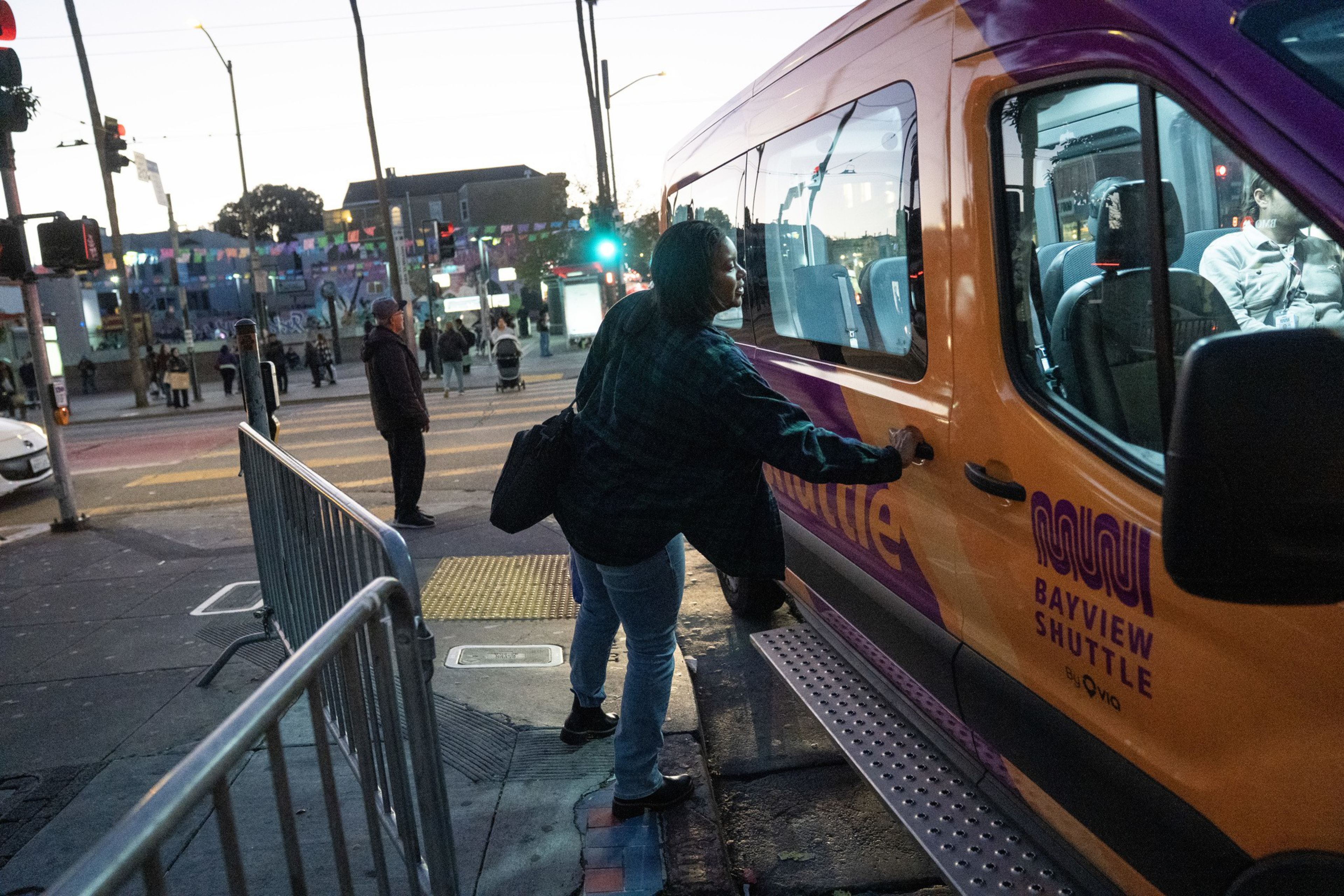 A person reaches to open the door of an orange Bayview Shuttle van parked by a street corner at dusk, with others walking in the background.