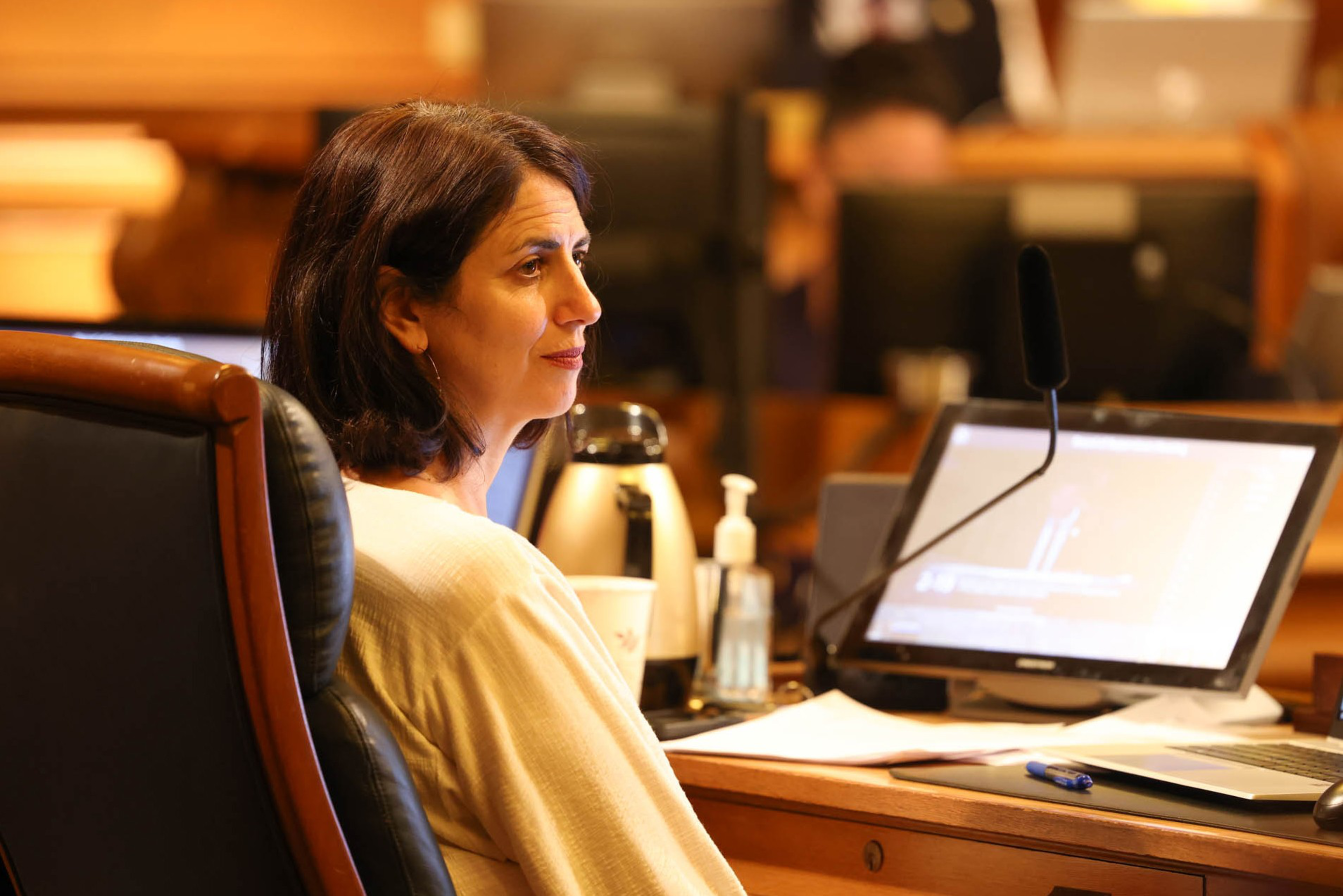 A side profile of a woman in a light sweater sitting in front of a dais with an open laptop.