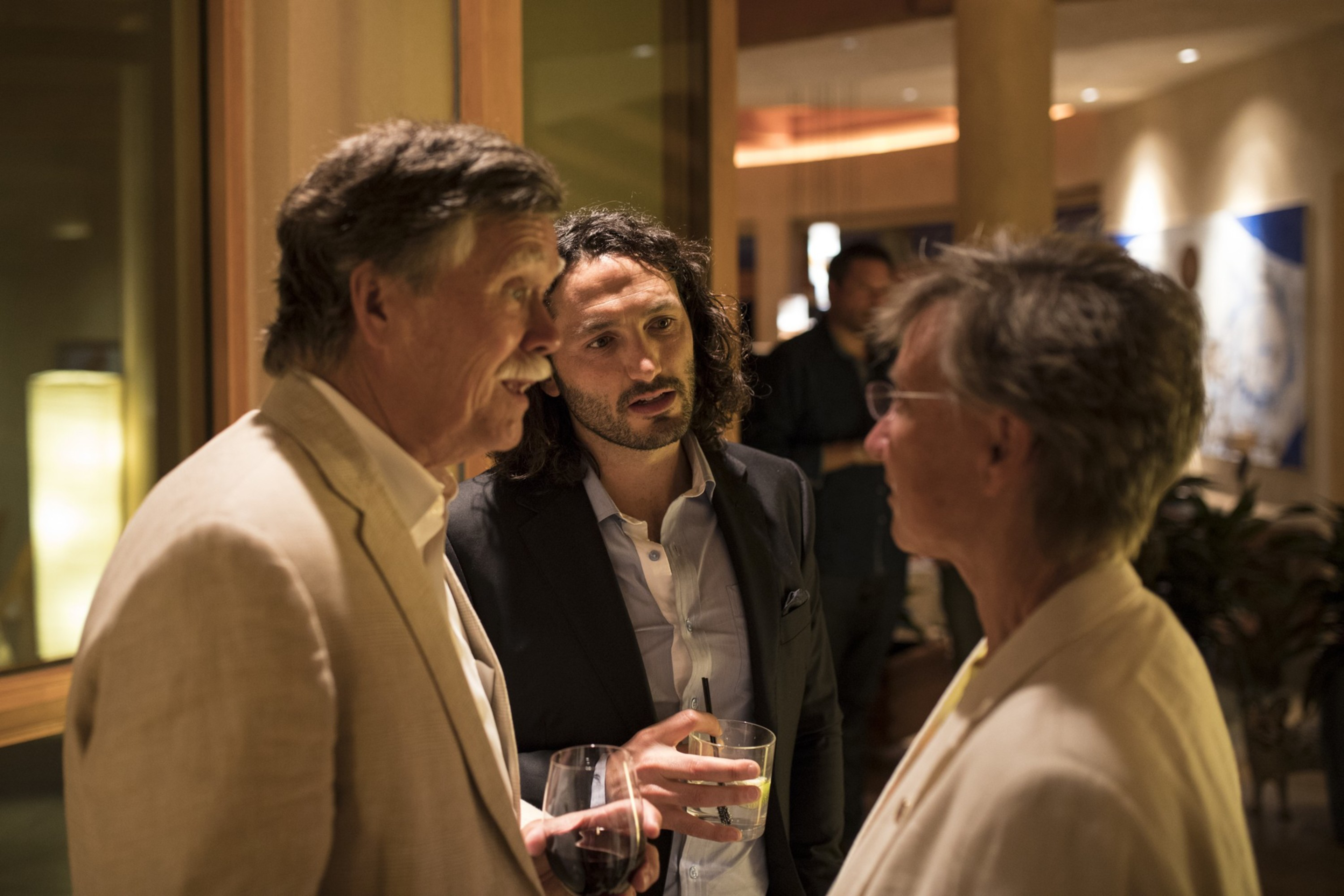 Three people in conversation at an indoor social event, two men and one woman, holding drinks and dressed in business casual attire.