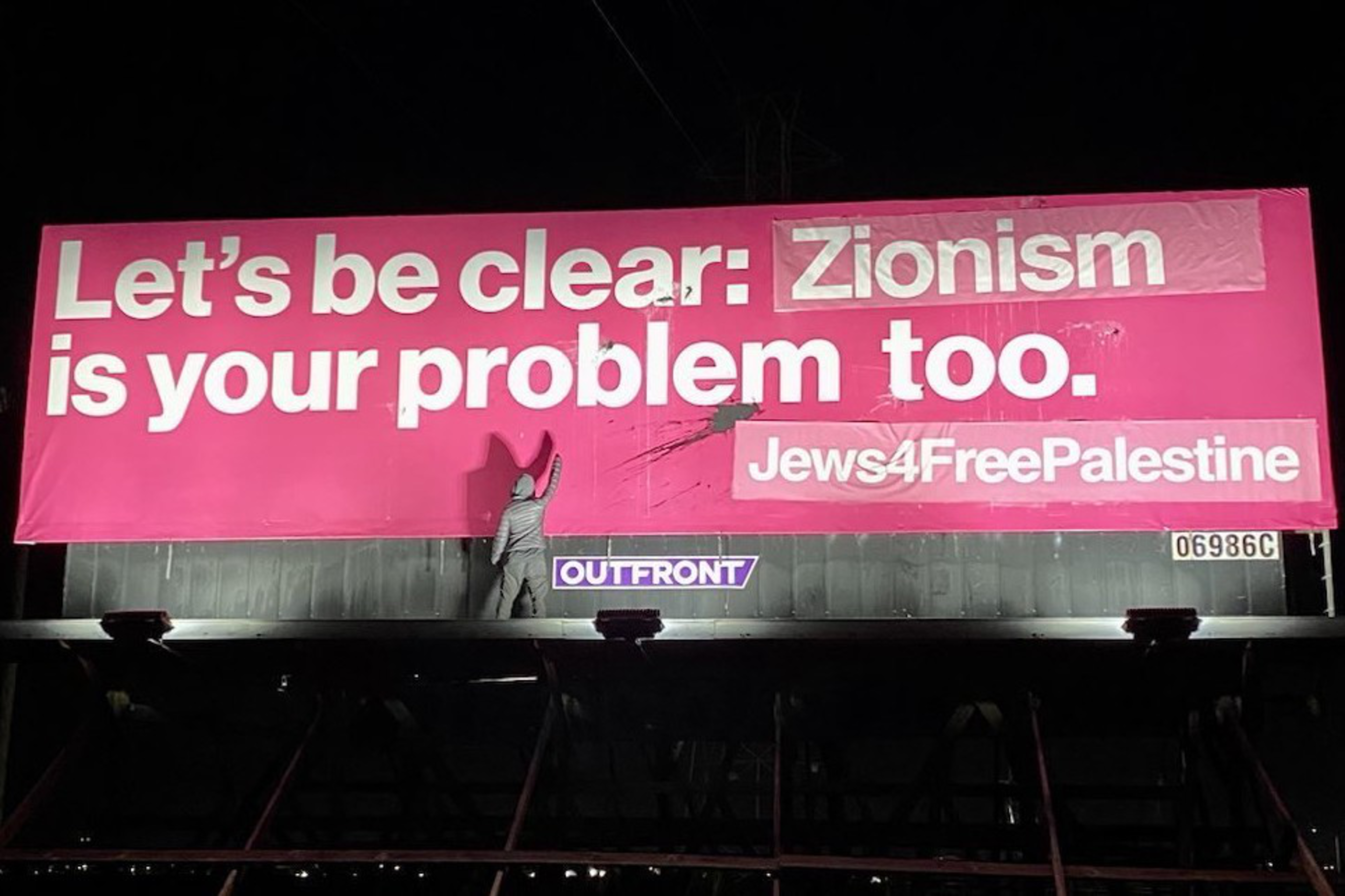 A person raises their fist while standing below a billboard that reads “Lets be clear: Zionism is your problem too. Jews4FreePalestine” in the evening.