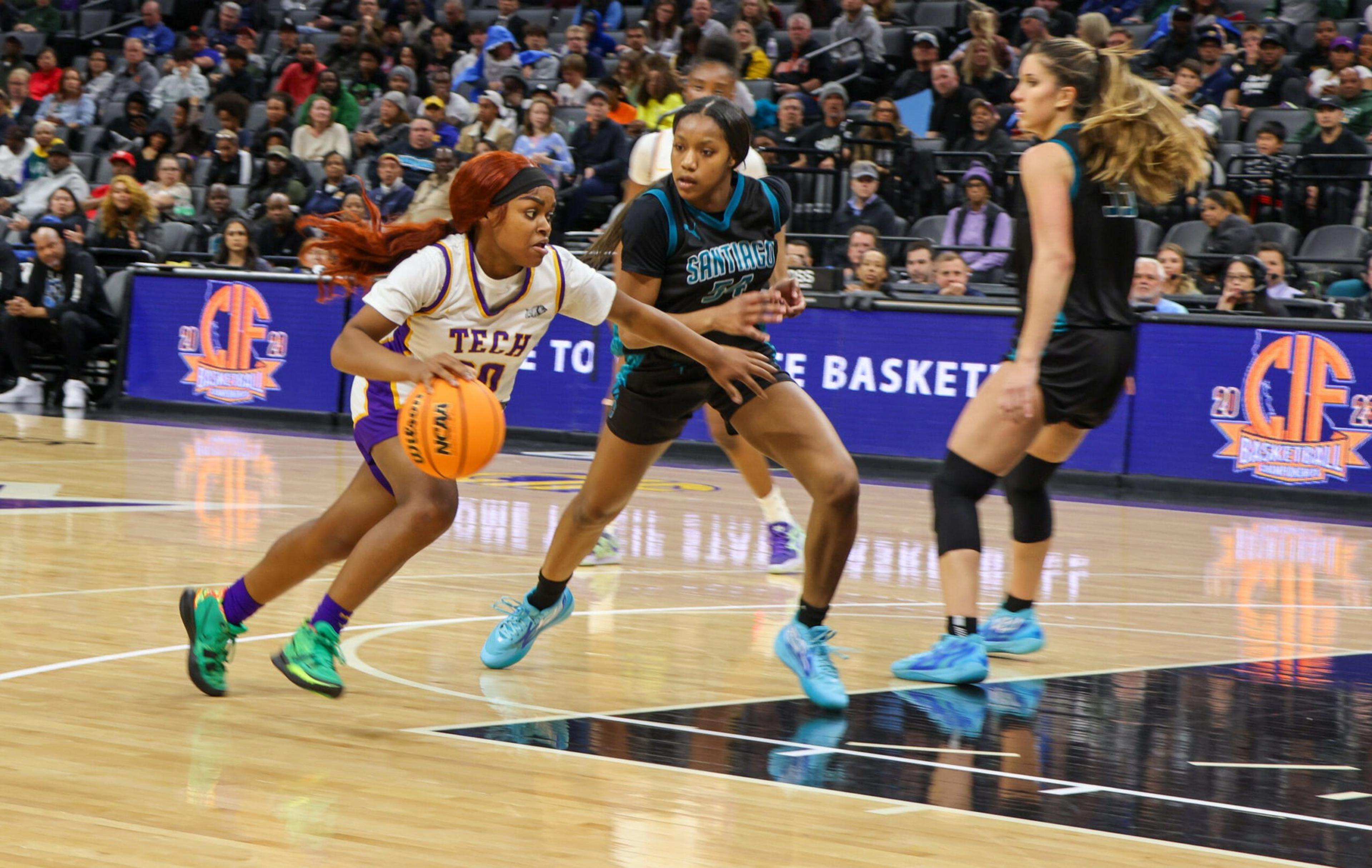 A female basketball player in a white and purple uniform dribbles past two defenders in black and teal uniforms on a crowded court.