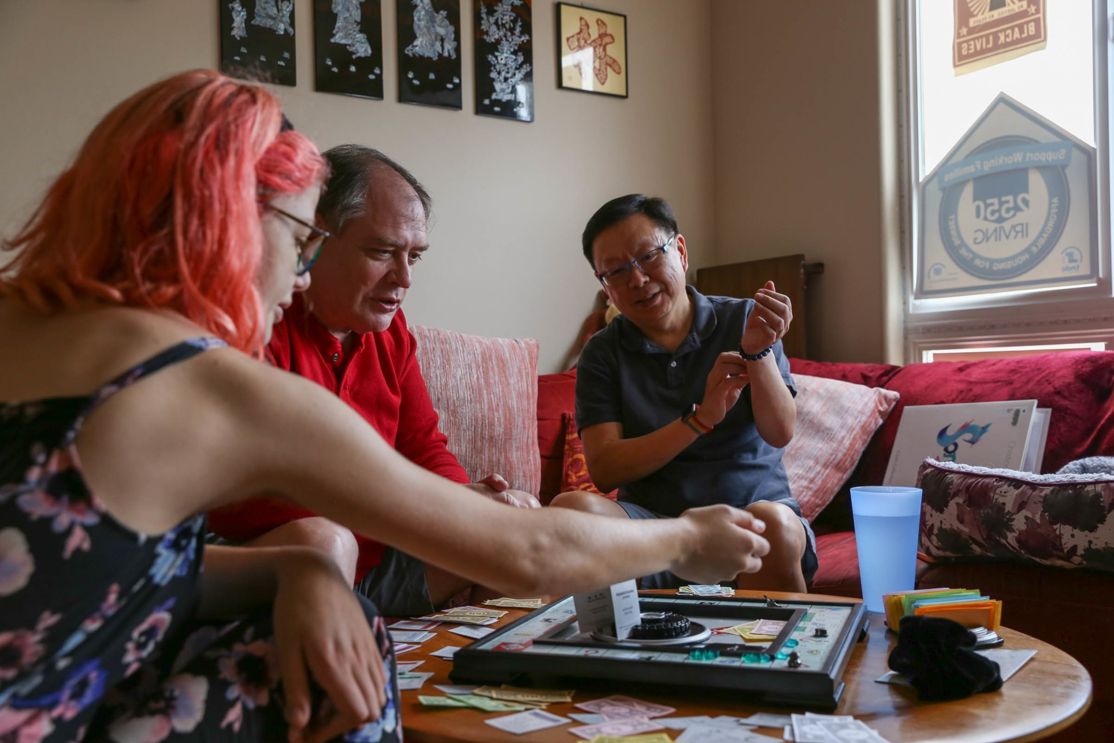 Three people sit on a couch and chair around a table, playing Monopoly with money and cards spread out, engaged and focused on the game.