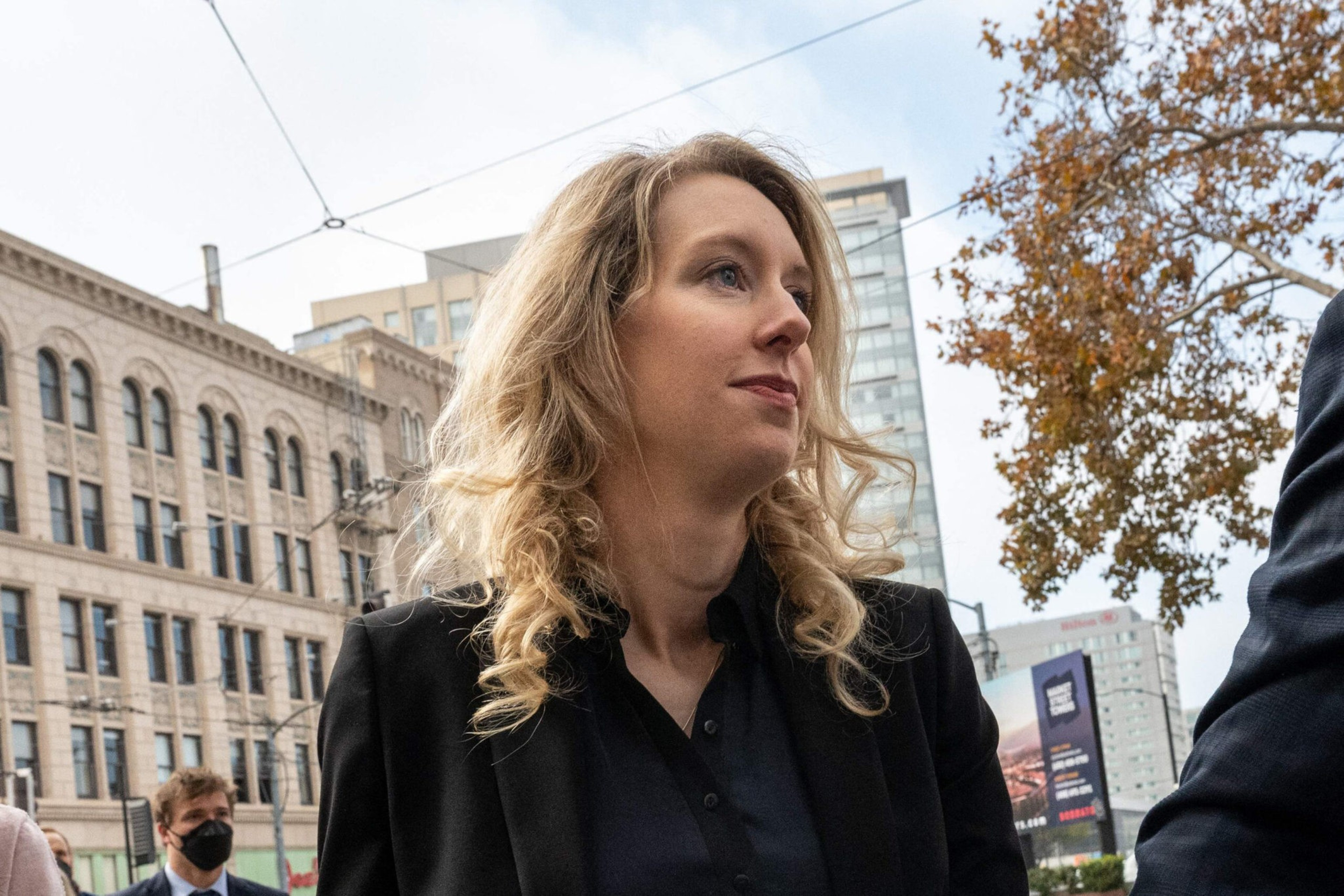 A blonde woman with curly hair wearing a black blazer looks slightly upward, with a cityscape and a tree with autumn leaves behind her.
