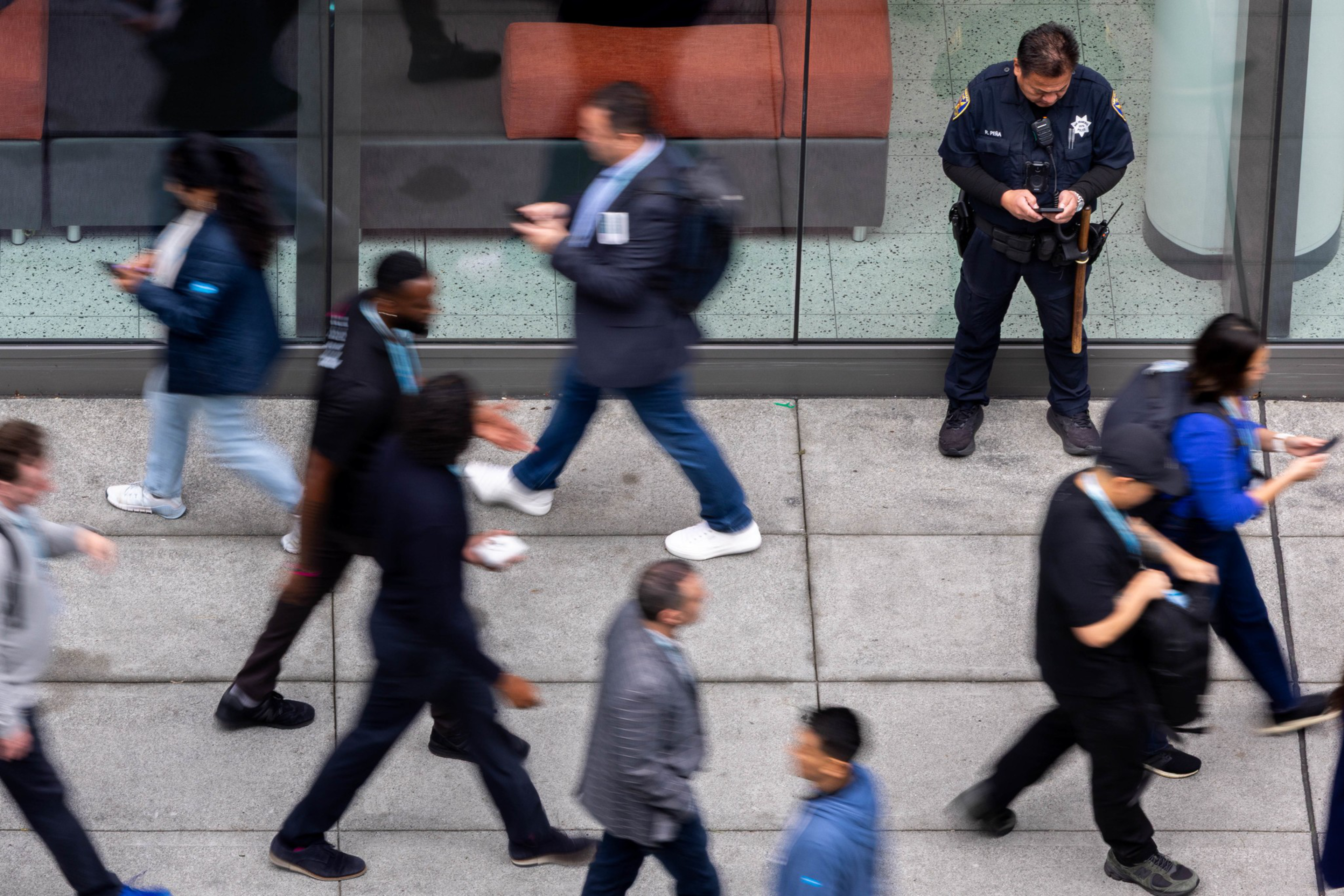 A police officer stands still against a wall while blurred pedestrians walk quickly past on a busy sidewalk.