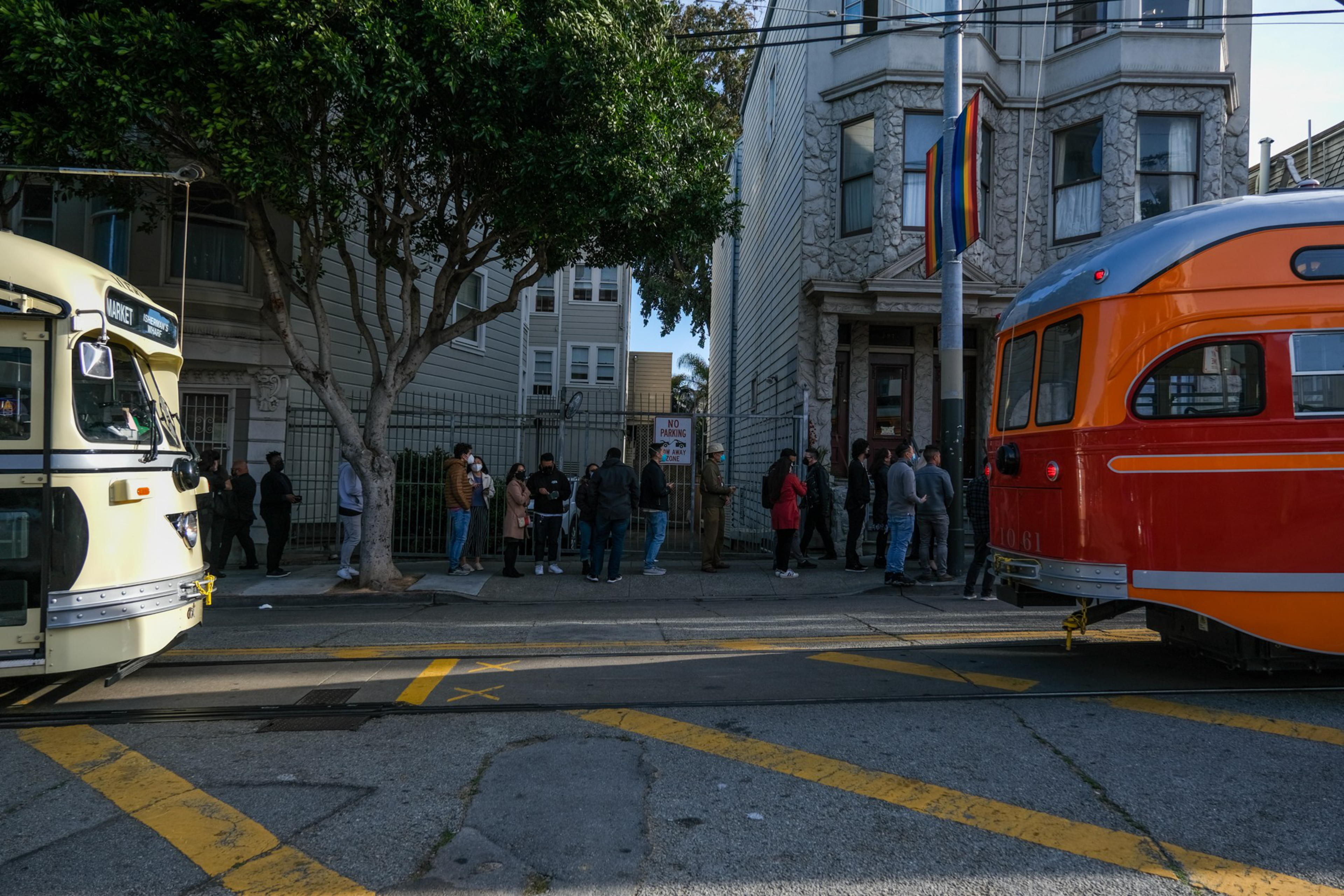 A diverse group of people stands in line on a sidewalk between an orange vintage streetcar and a cream-colored vintage trolley.