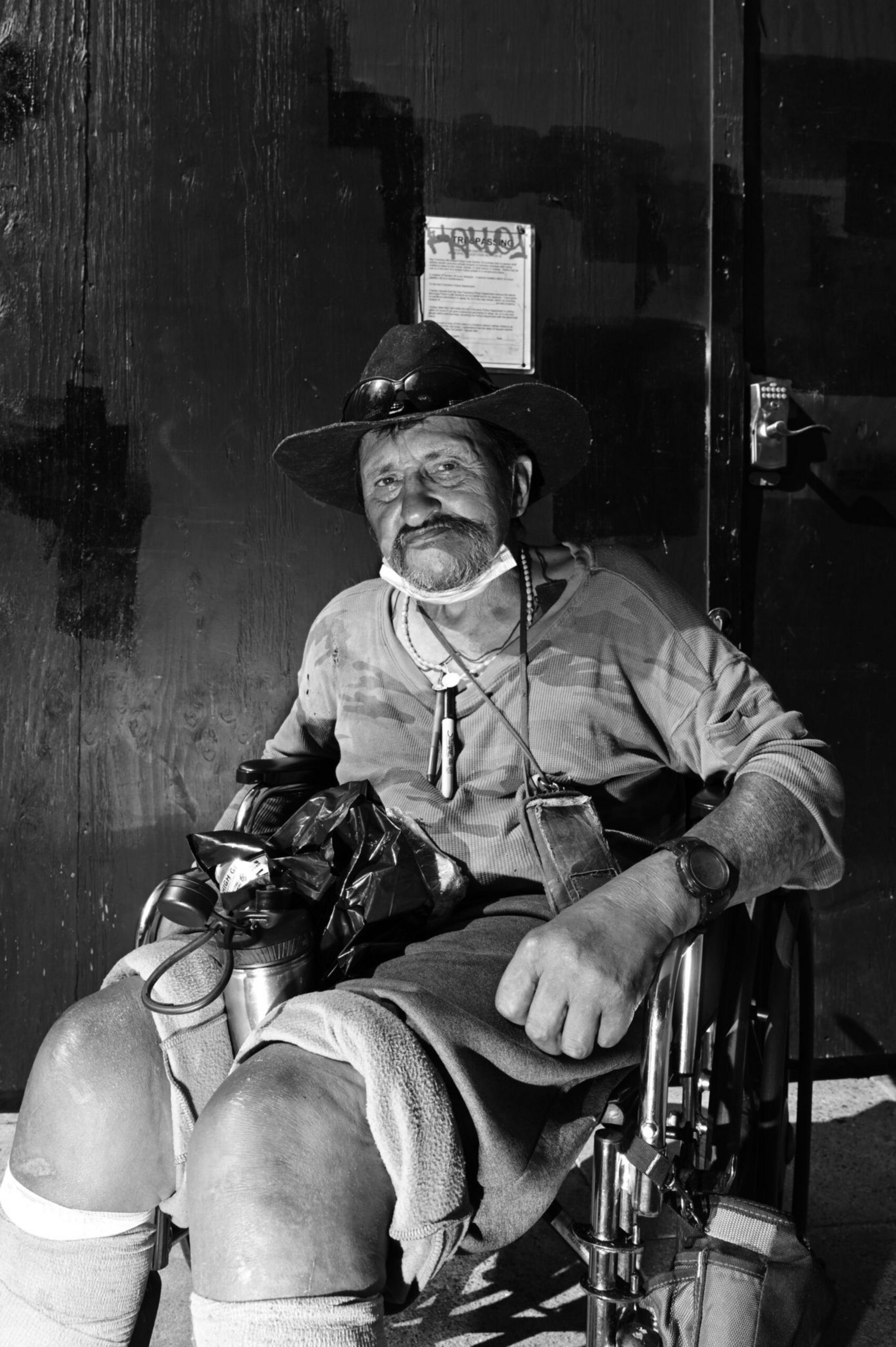 An older man with a mustache sits in a wheelchair, wearing a wide-brimmed hat, camo shirt, and a mask pulled below his chin.