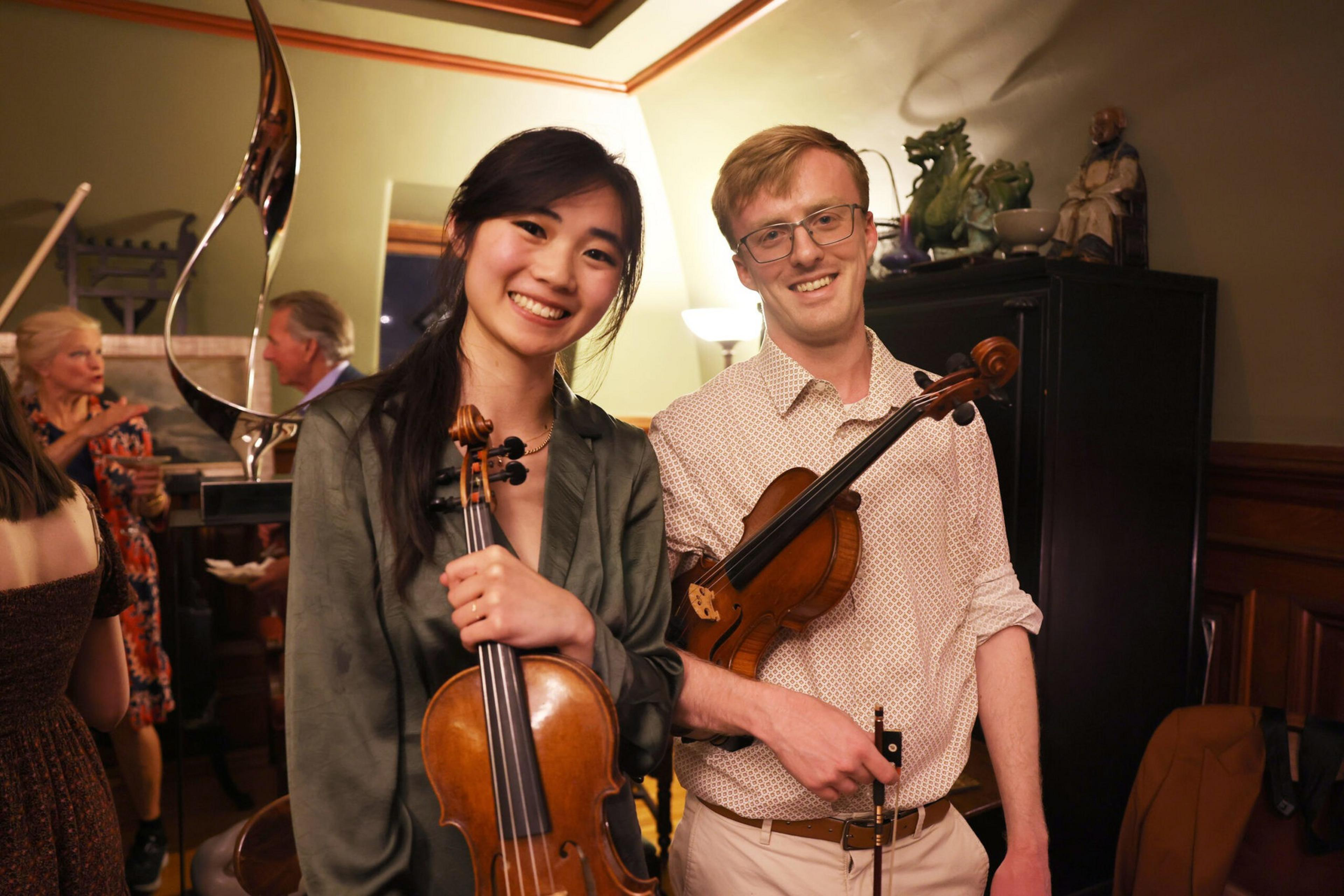 A smiling Asian woman and a Caucasian man each hold violins in a warmly lit room with other people and decorative items in the background.