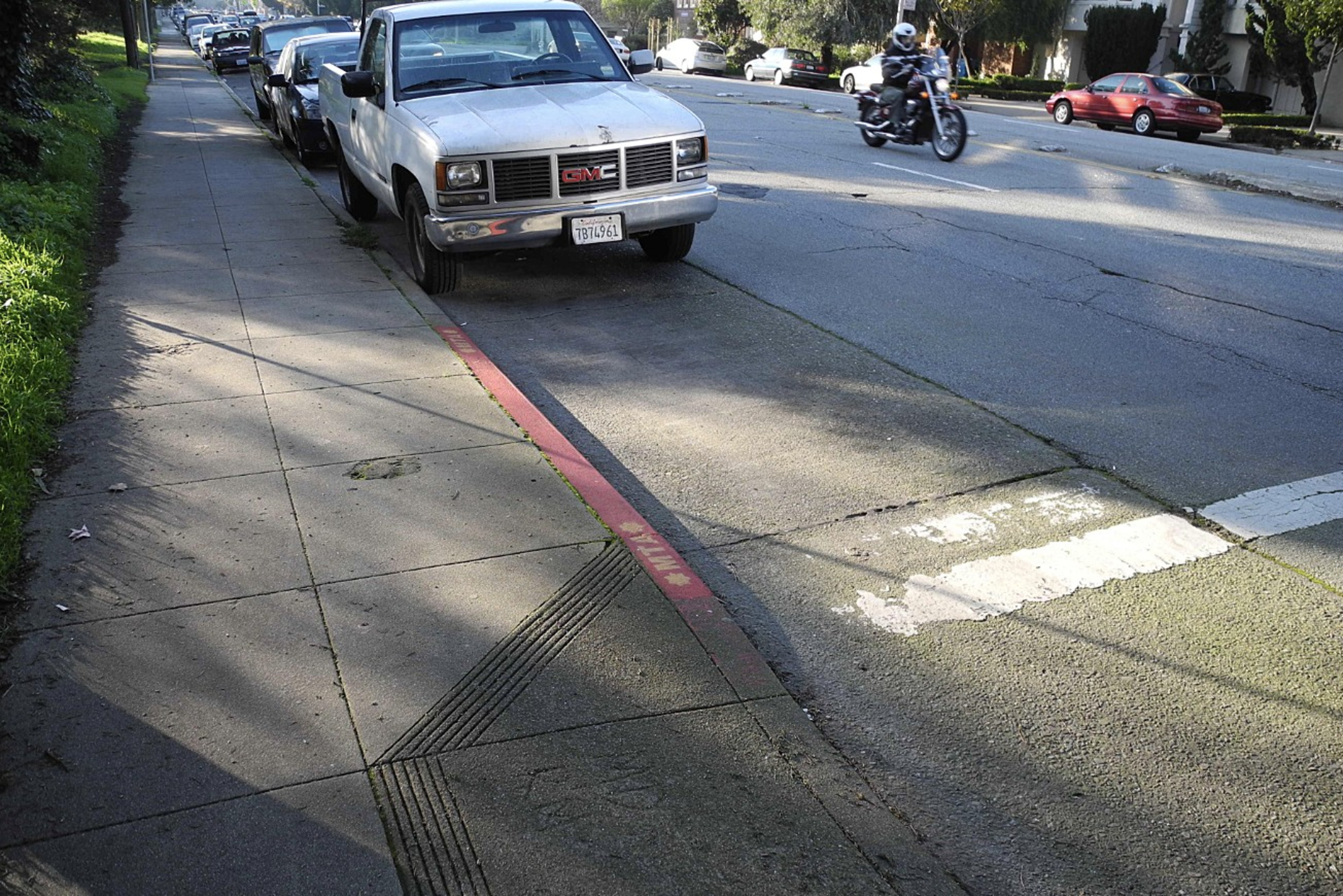 A truck is parked on a sidewalk next to a street where a motorcycle passes by. There's a red curb and greenery along the walkway.