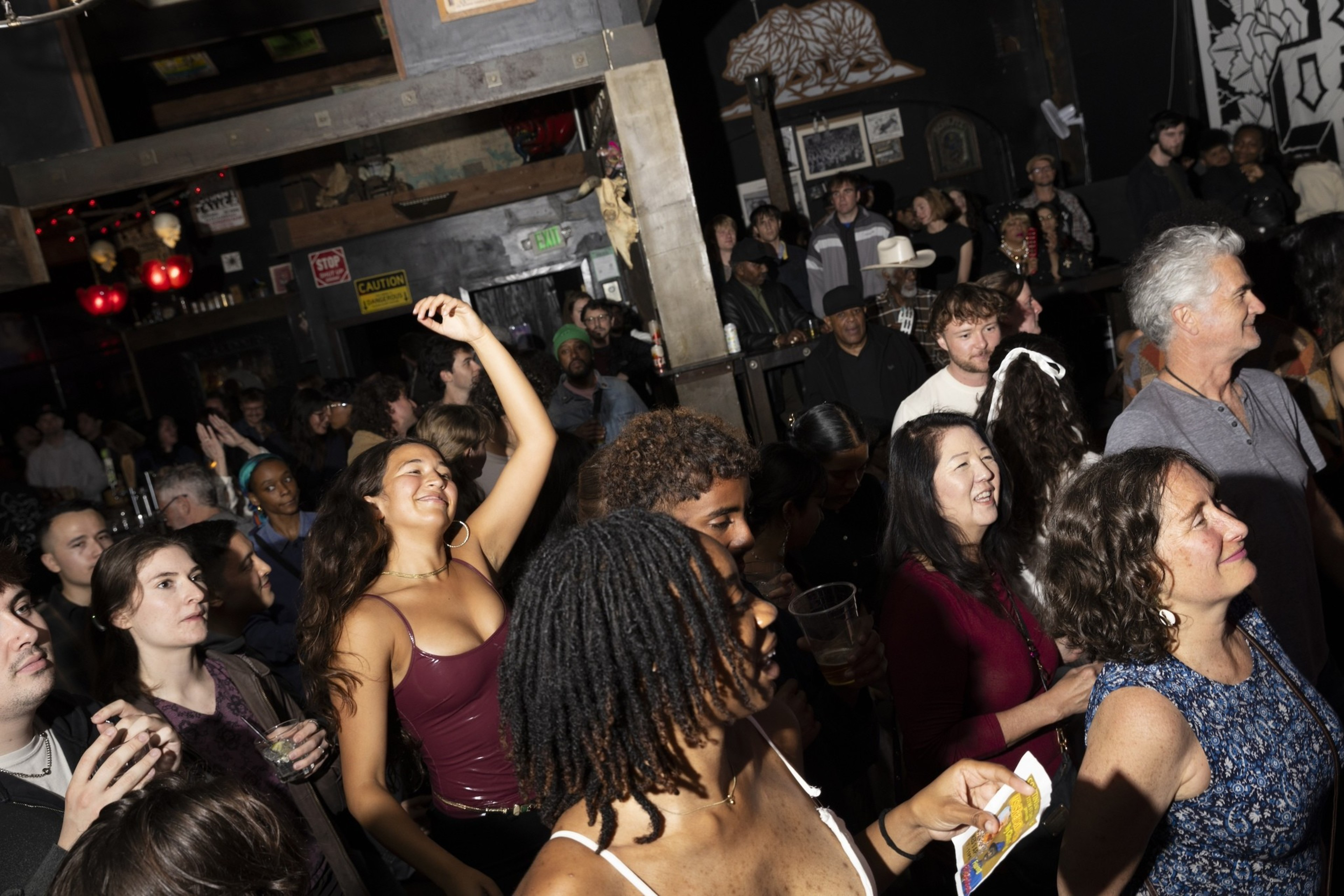 A lively crowd at a dimly lit venue, with people smiling, drinking, and dancing. Decor includes red lights and various wall art, creating a festive atmosphere.