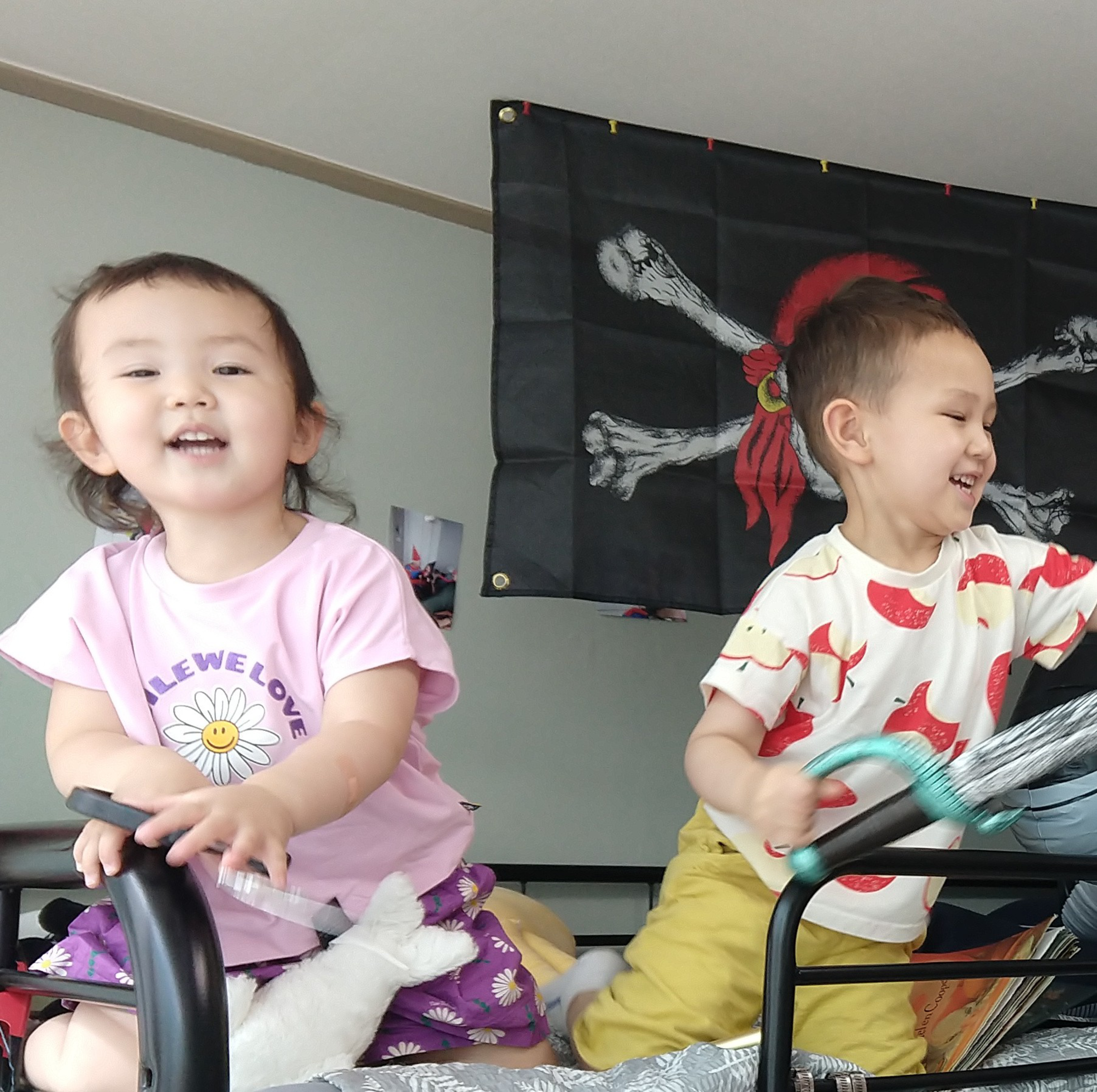 Two young children happily play on a bed, one holding a black rail and the other holding a toy sword, with a pirate flag hanging on the wall behind them.
