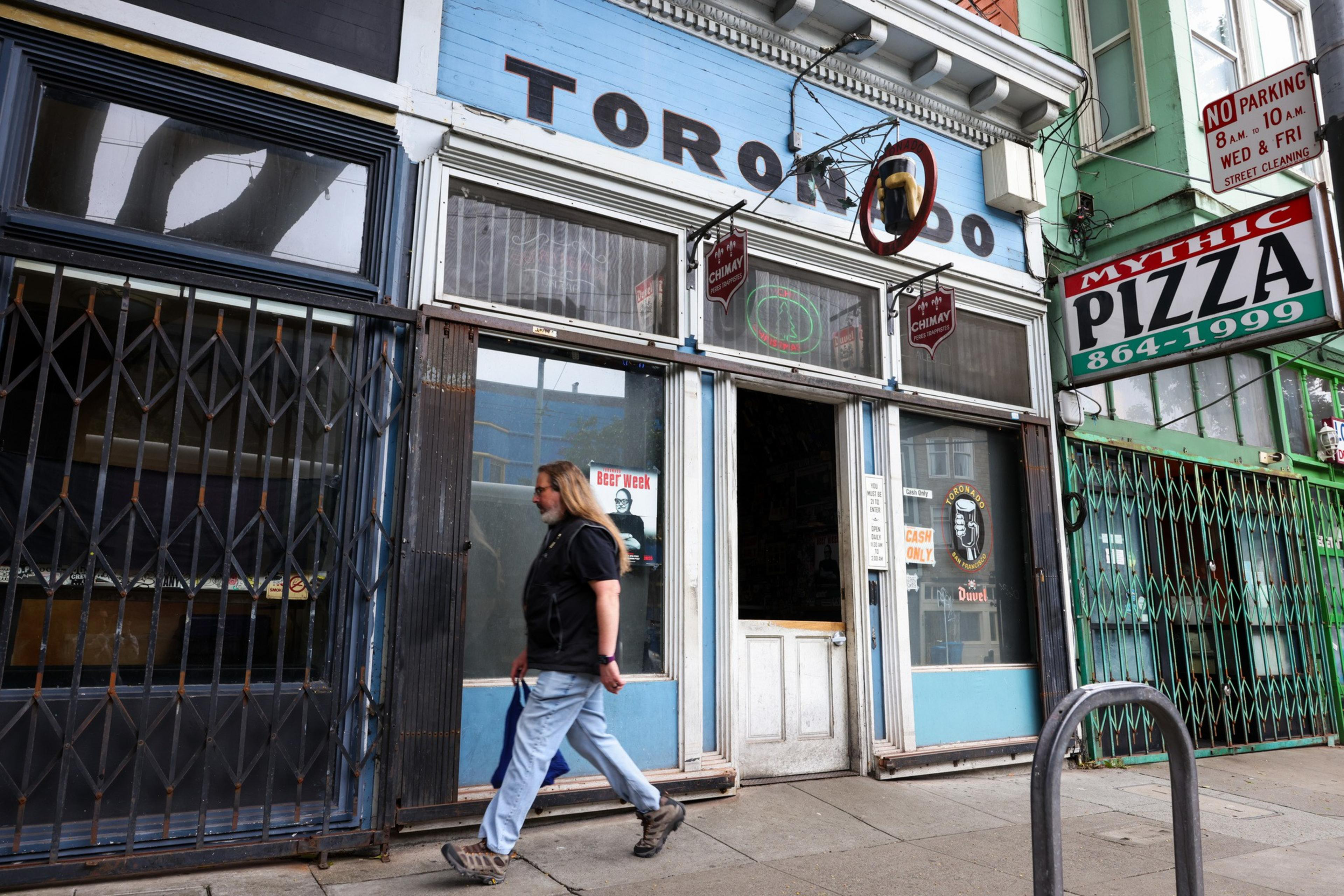 A person walks past a storefront with a sign that reads "TORONADO" next to a "MYTHIC PIZZA" sign. The door is open, revealing posters and signs inside.