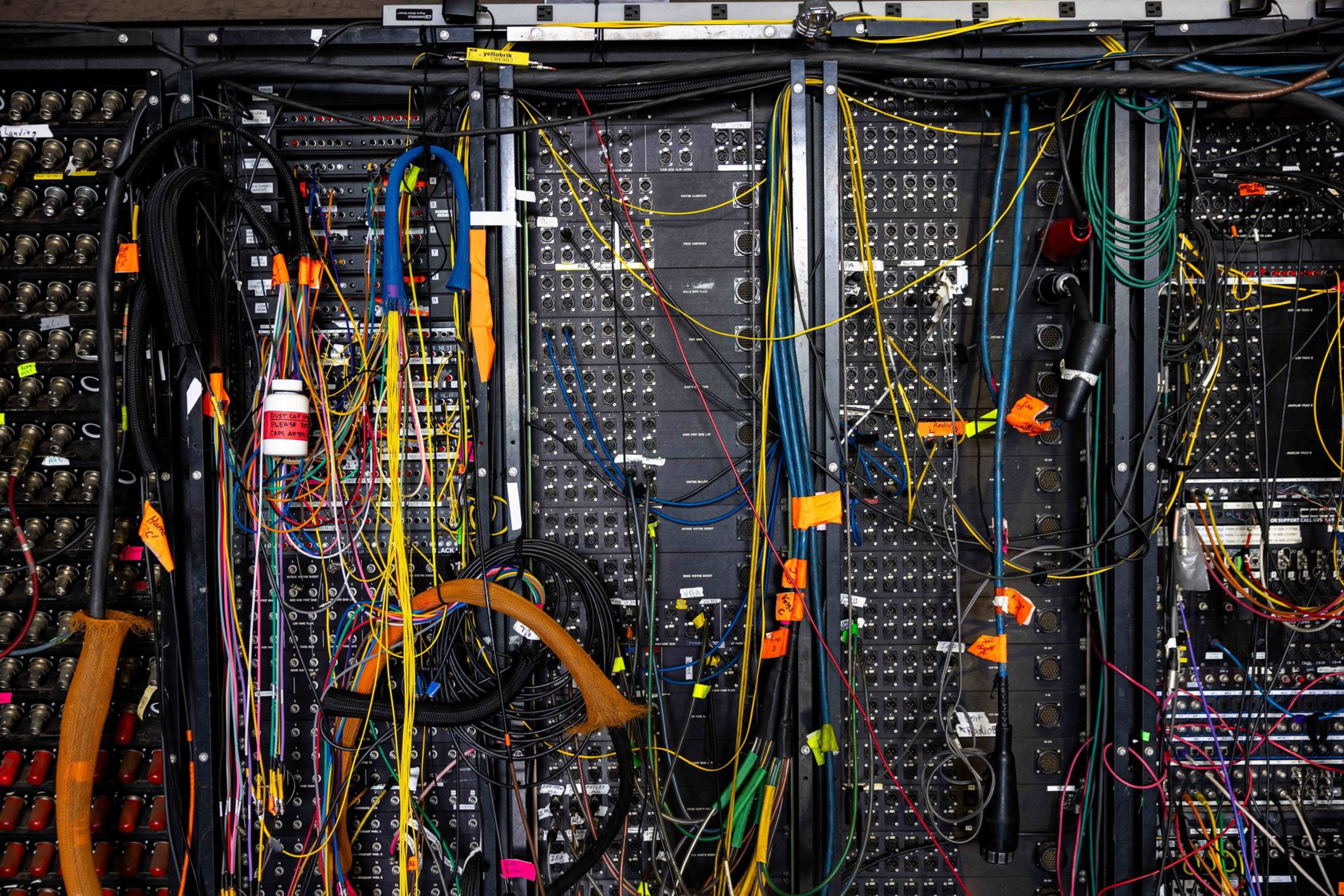 A complex wall of audio or electronic equipment with numerous cables of different colors connected and hanging, some labeled with orange tags.