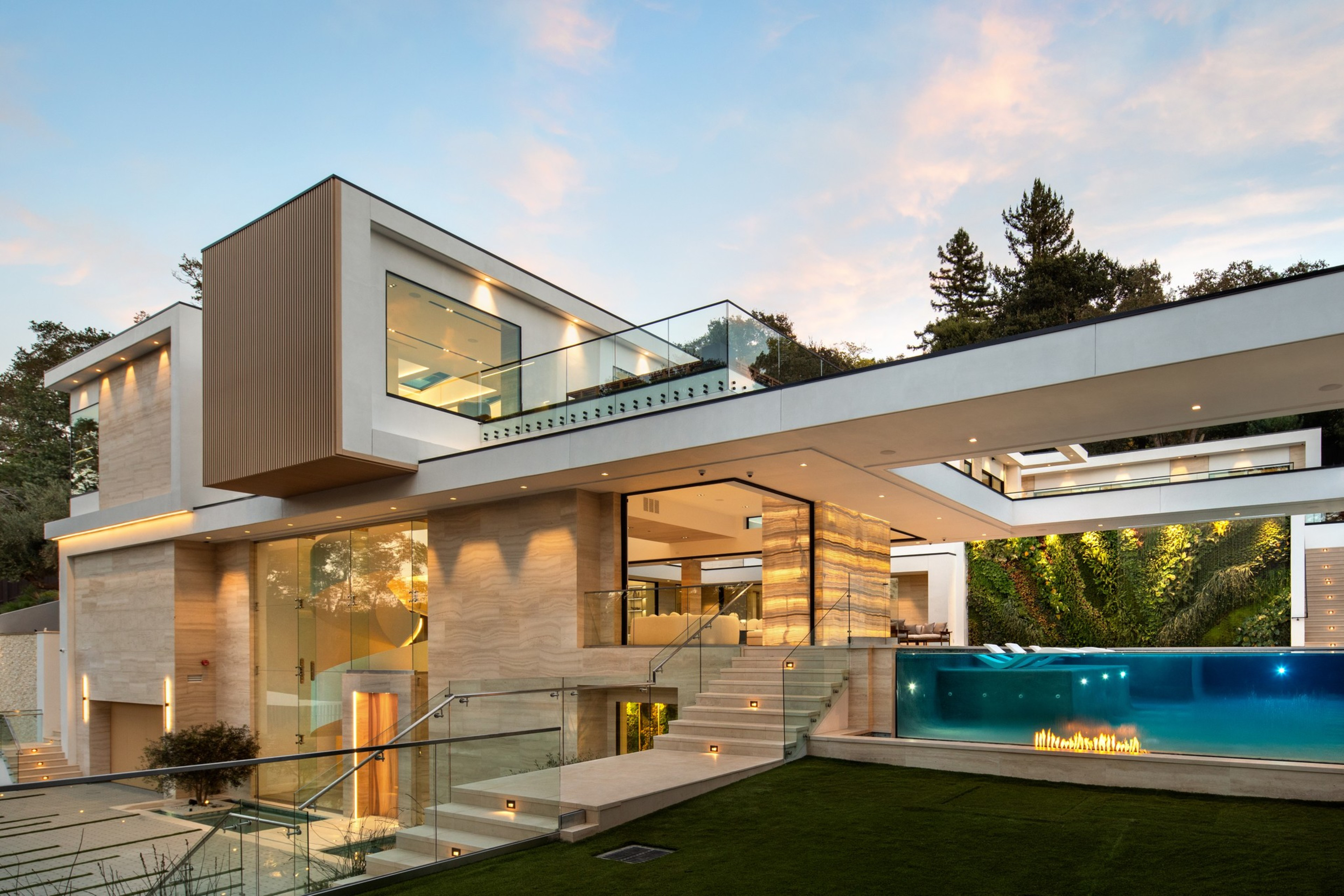 A modern multi-level house with large glass windows, a rooftop terrace, an illuminated pool with a fire feature, and stairs surrounded by greenery at dusk.